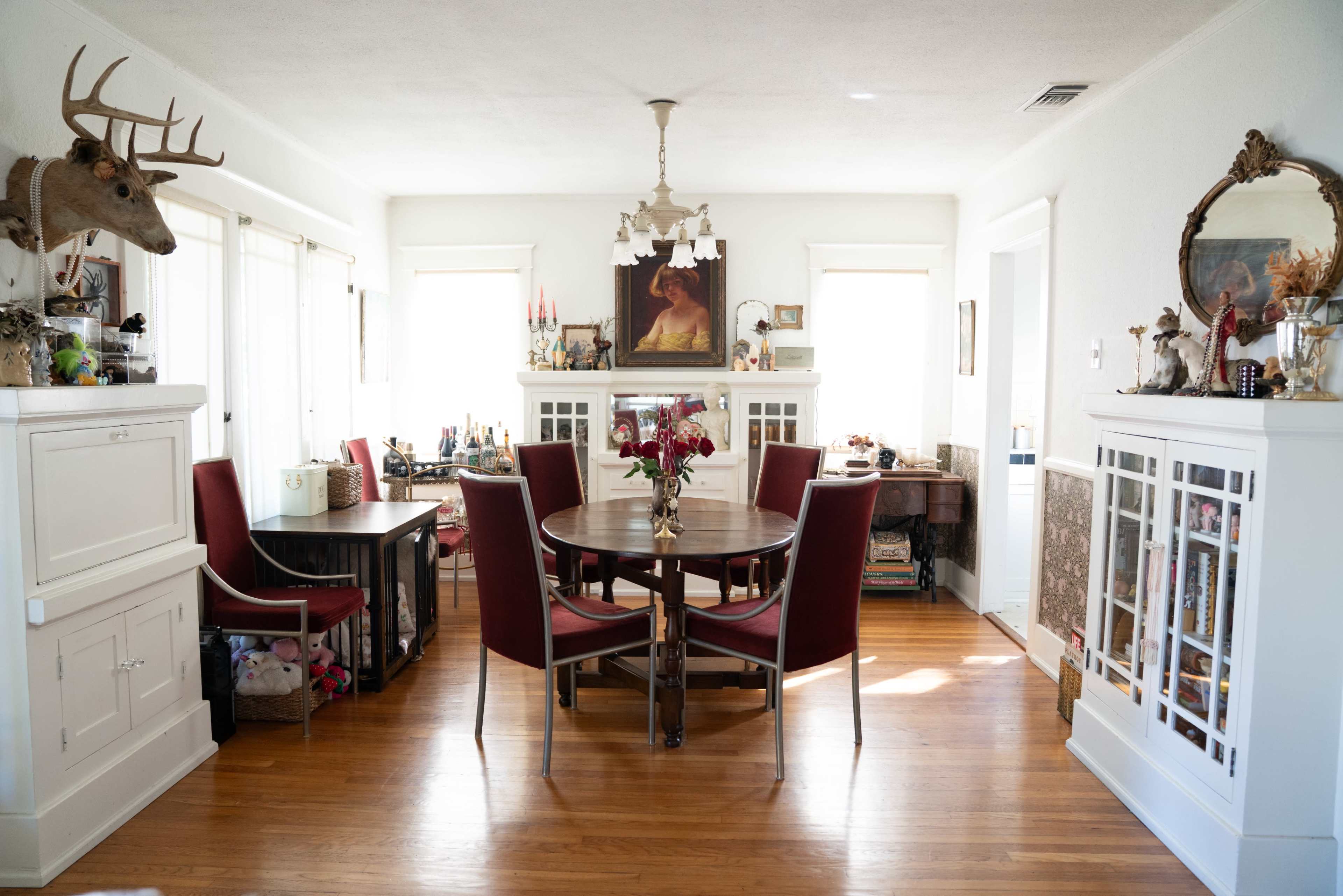 The image shows a cozy dining room with a round table surrounded by red chairs, a decorative chandelier, and various items displayed on shelves and walls.