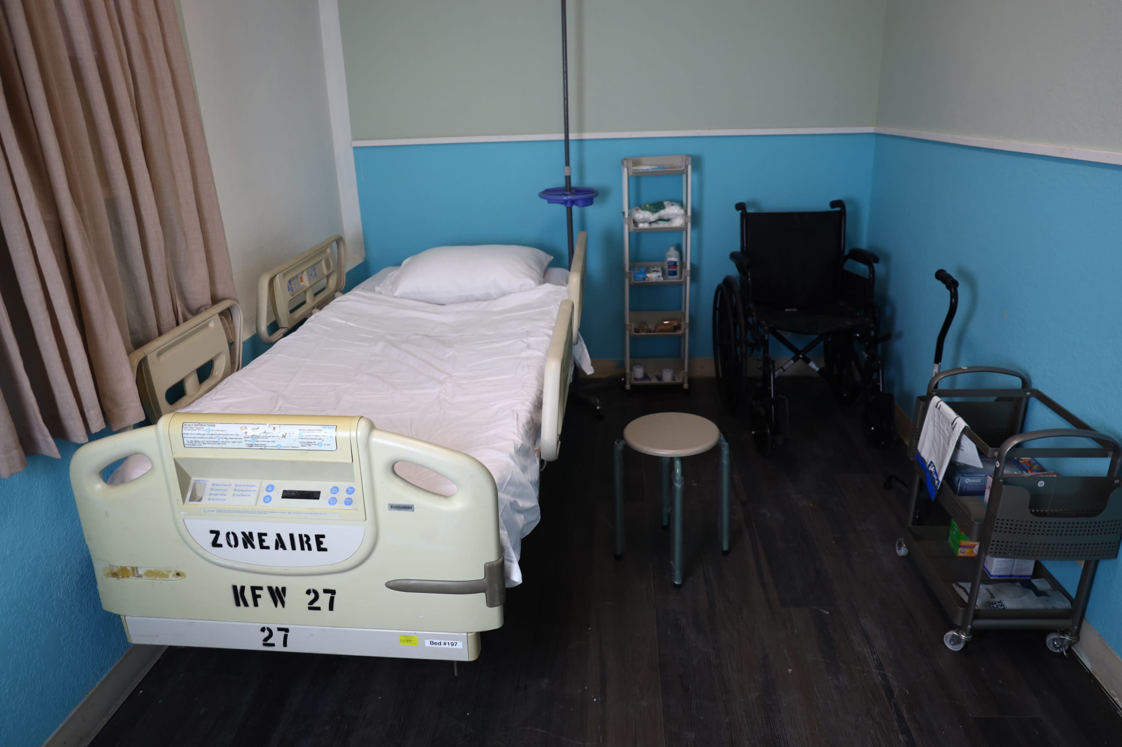 The image shows a small hospital room with a bed, a wheelchair, a stool, and medical equipment on carts against a blue wall.