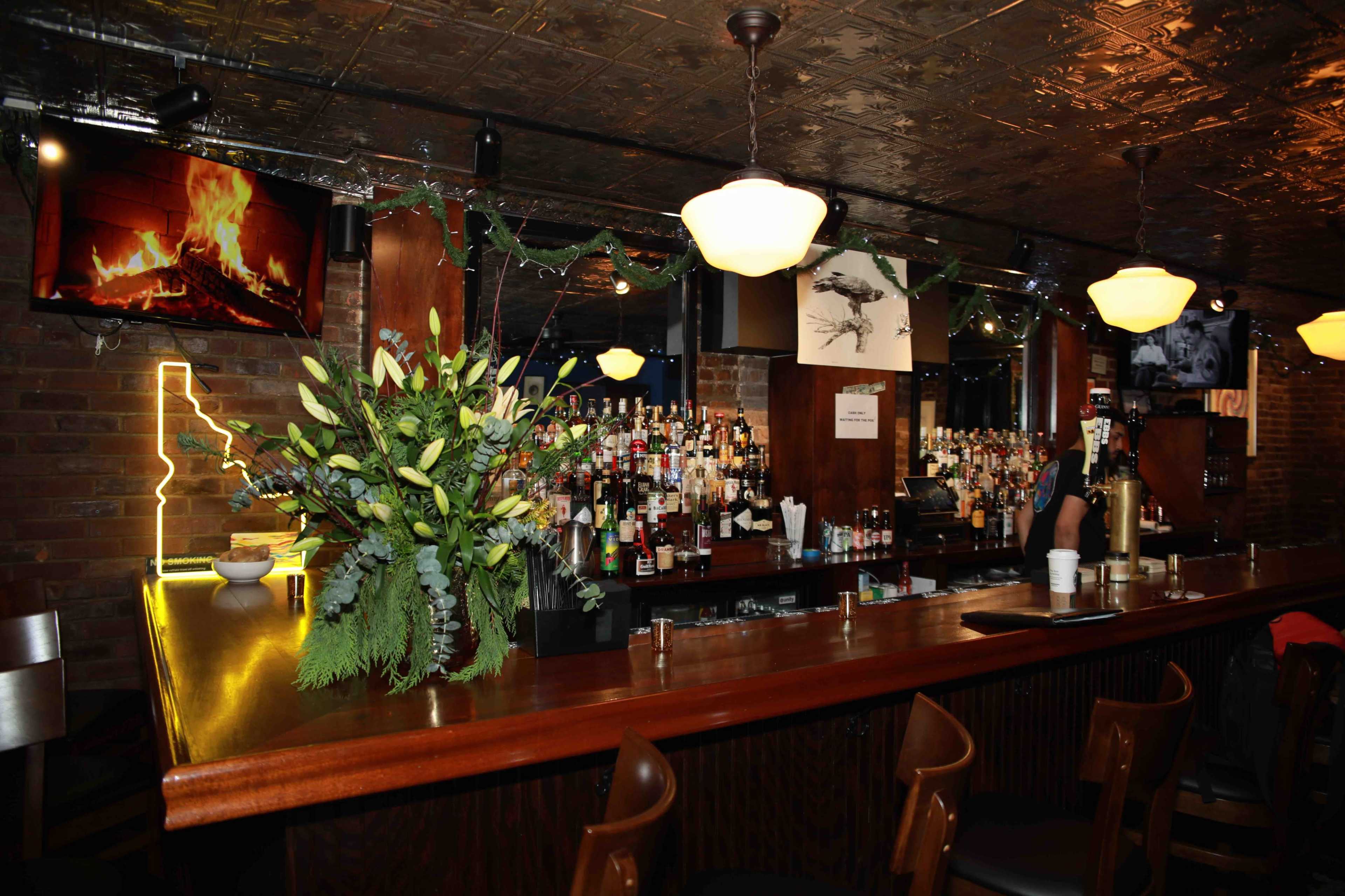 A bar with a wooden counter, a variety of alcoholic beverages on display, and decorations including a fireplace on a screen and a floral arrangement.
