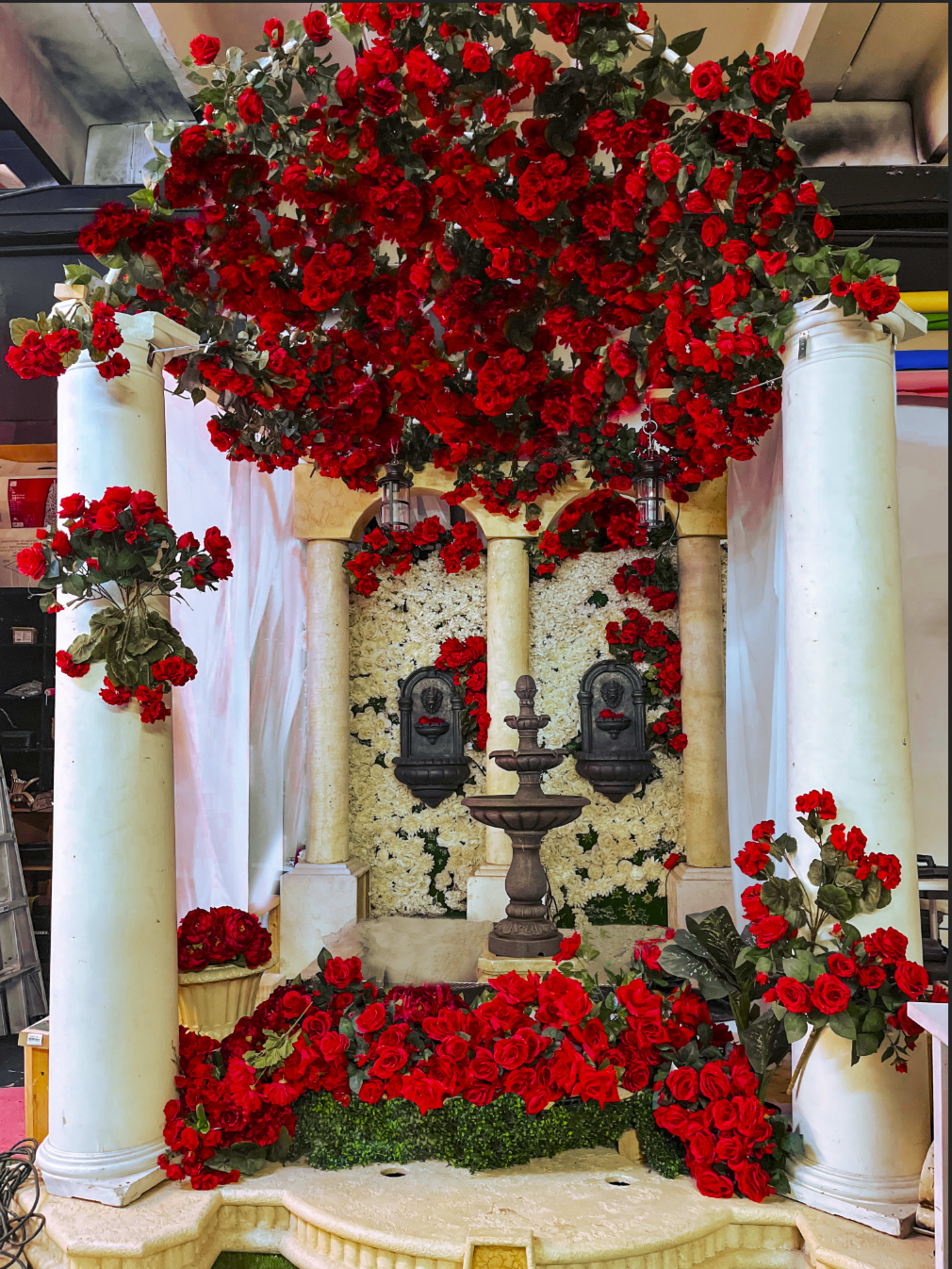 The image shows a decorative fountain surrounded by red roses, flanked by two columns, with additional rose arrangements above and on the sides.