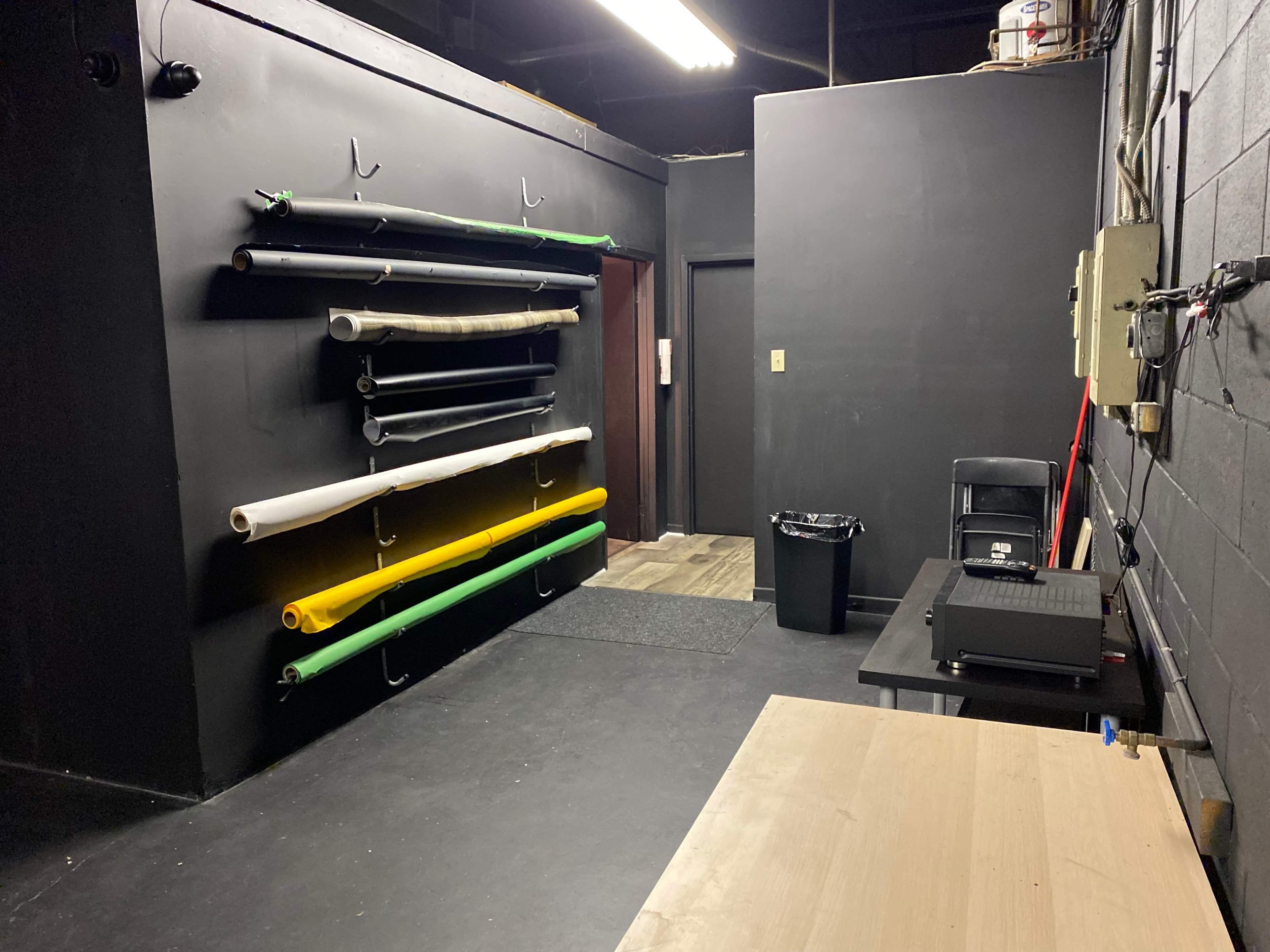 The image shows a dimly lit room with black walls, featuring a rack of colorful rolls of material on one wall and a table with a printer and chair on the opposite side.
