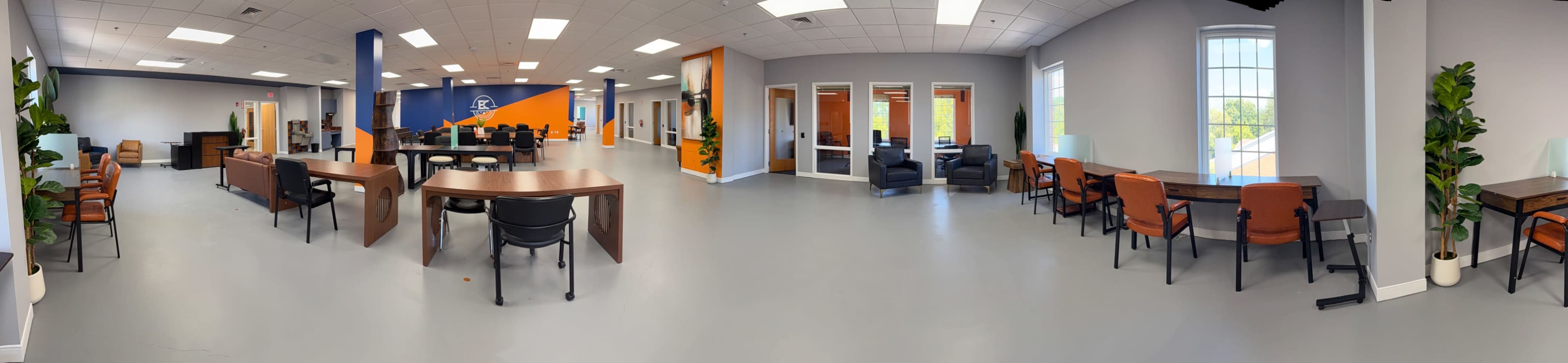 The image shows a spacious, modern office environment with various tables, chairs, and lounge areas, featuring a blue and orange accent wall.