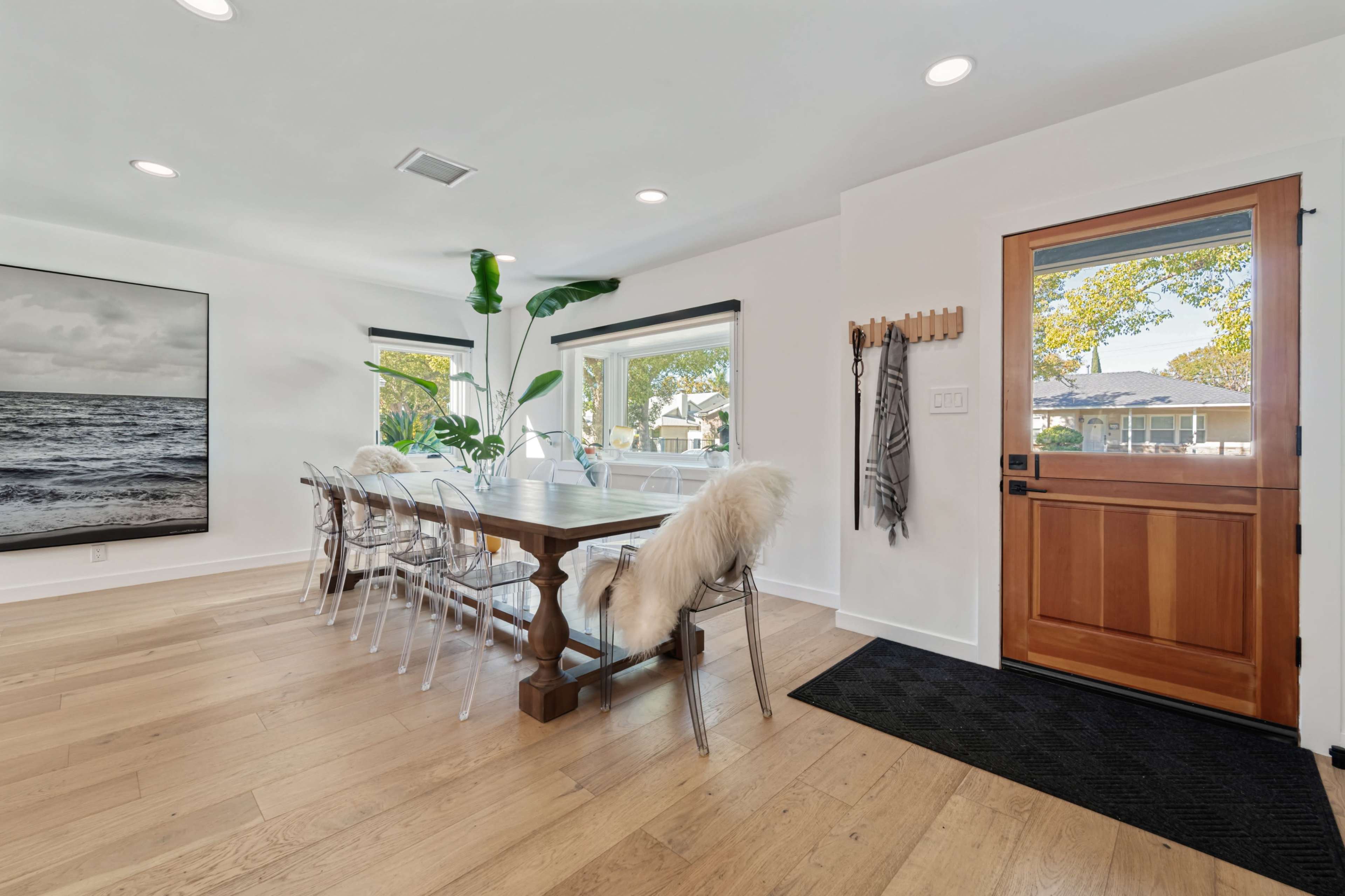 The image shows a modern dining area with a wooden table surrounded by transparent chairs, large windows, and a front door leading to an entrance.