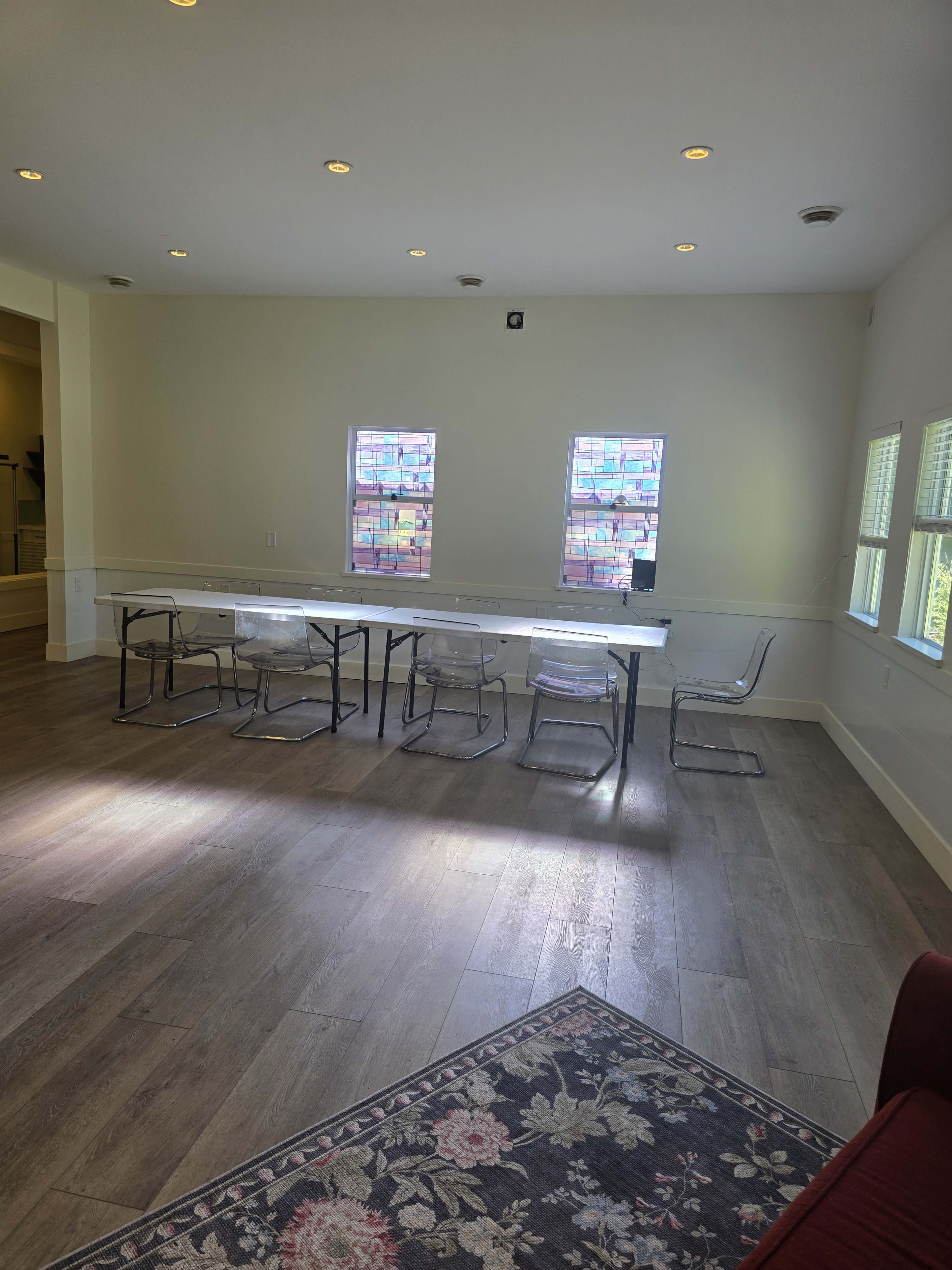 The image shows a spacious room with a long table surrounded by clear chairs, featuring two windows that let in natural light and a rug in the foreground.