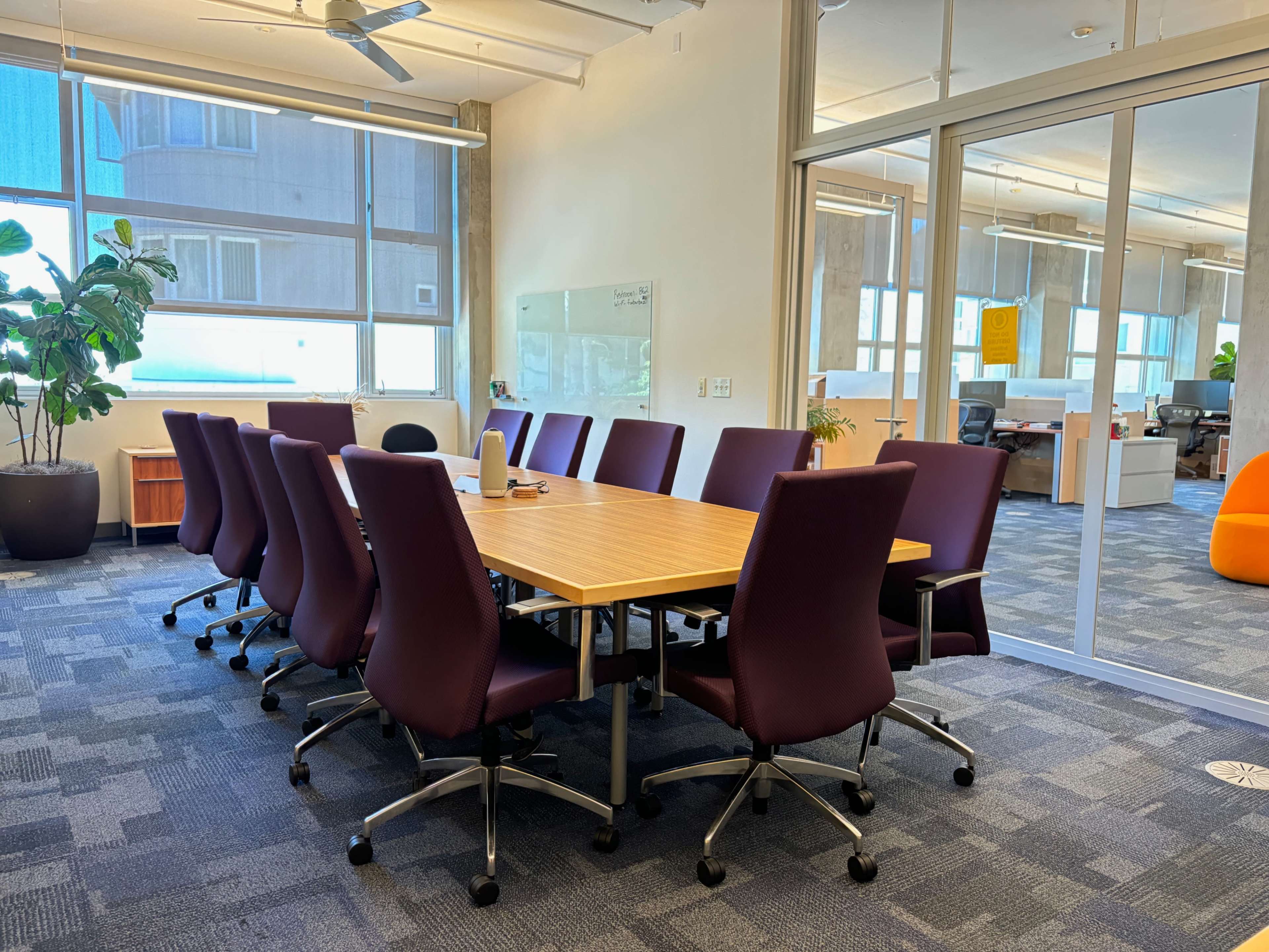 A conference room features a large wooden table surrounded by twelve purple chairs, with large windows allowing natural light.