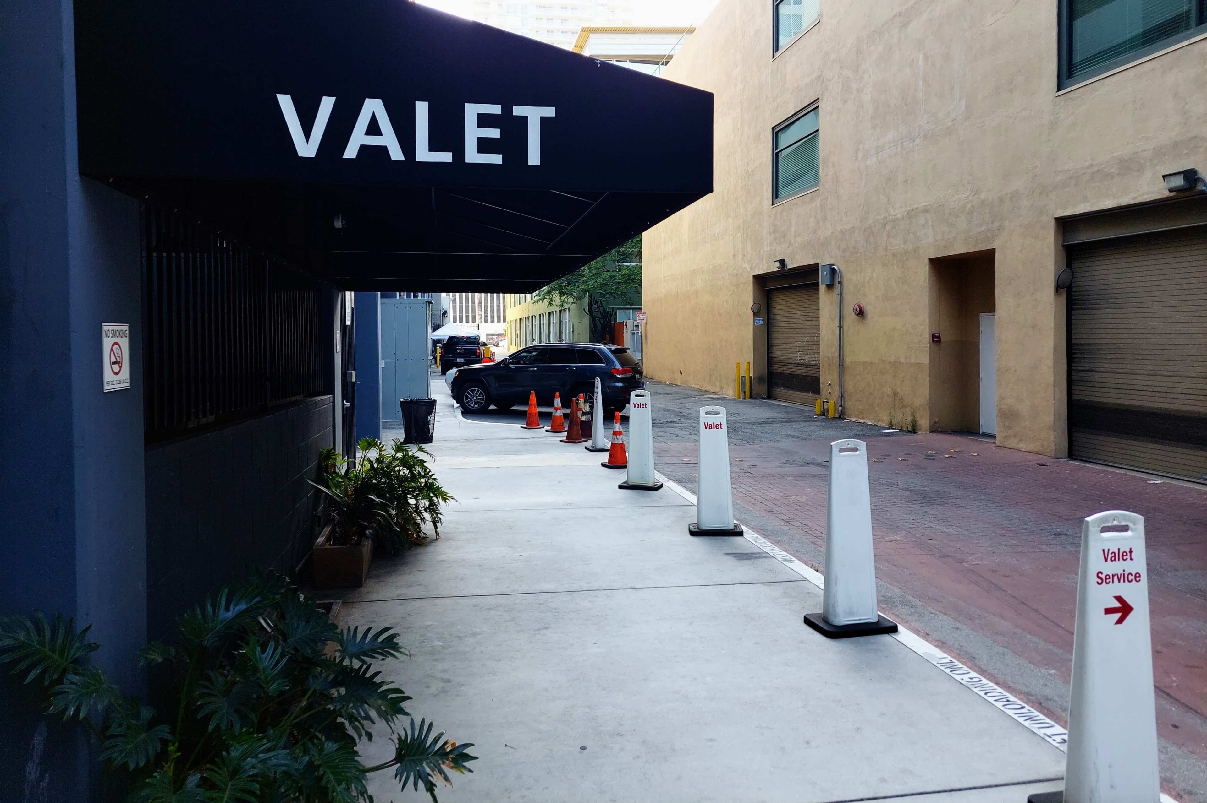 A valet area with a canopied entrance, marked by a sign, and lined with traffic cones and white poles, is situated beside a building and leads to a parking area.