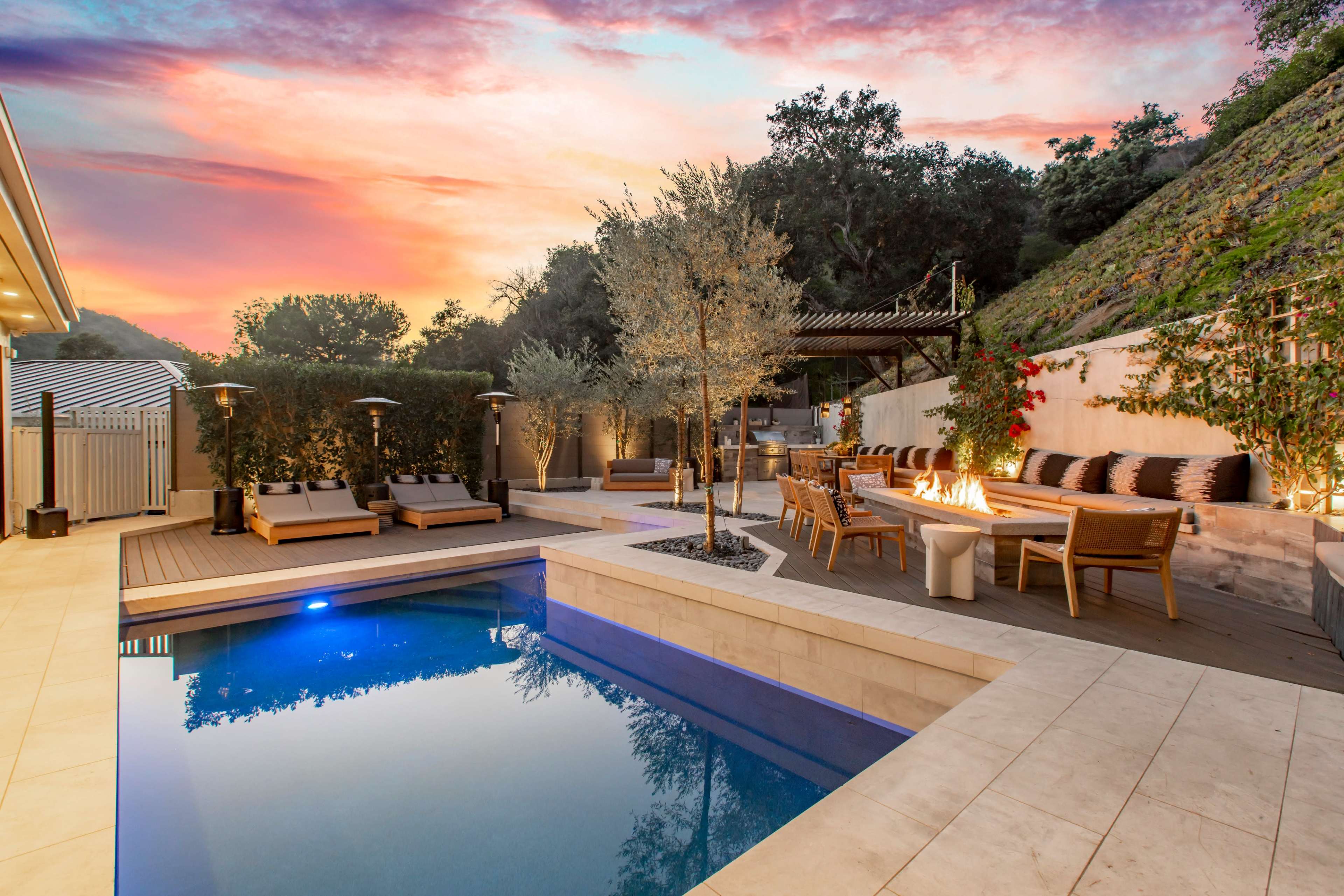 The image features a modern outdoor space with a swimming pool, surrounded by seating areas, a fire pit, and lush greenery, all under a colorful sunset sky.