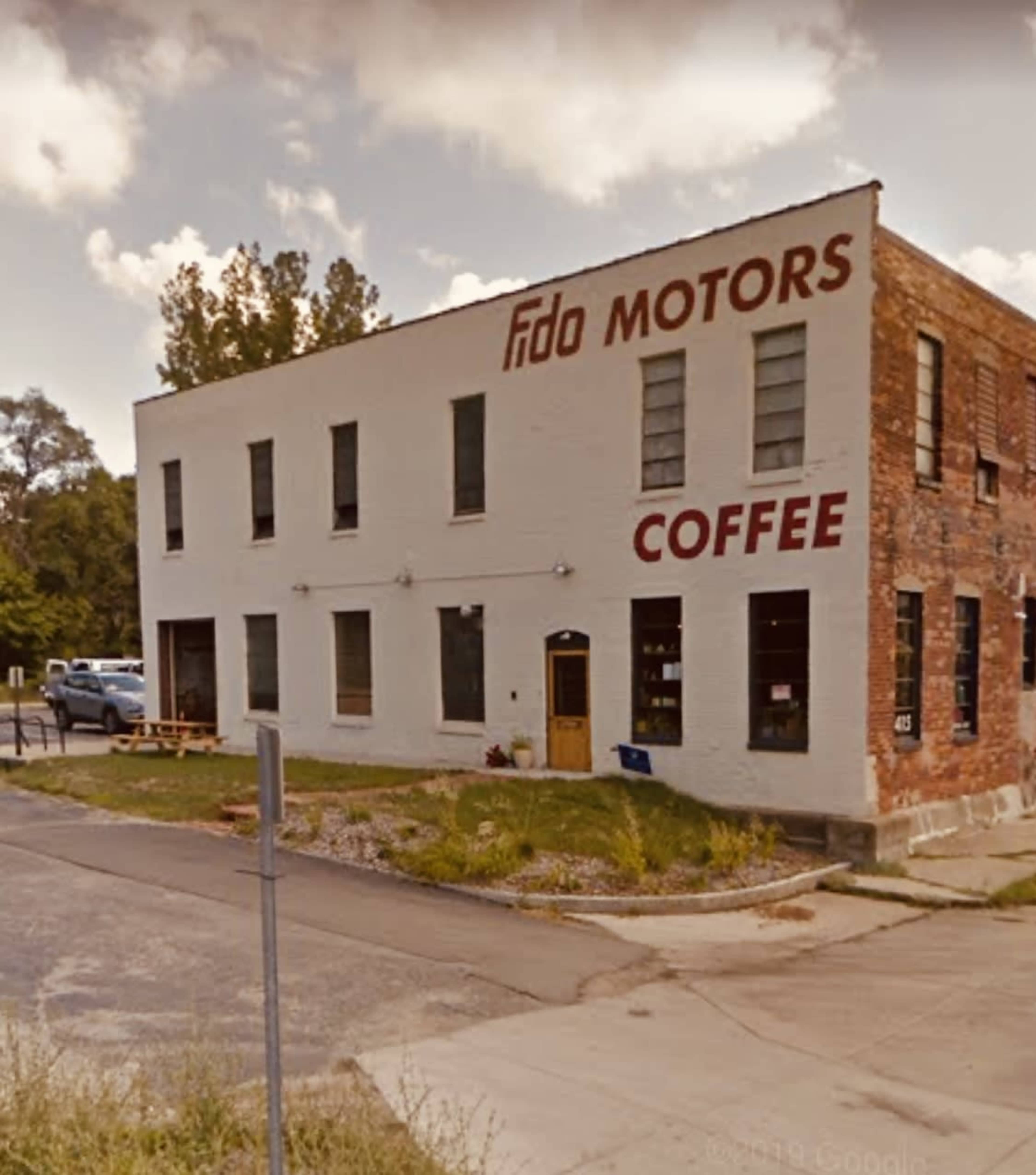 A two-story building featuring a sign that reads "Fido Motors" and "COFFEE," with a landscaped area in front and a parked vehicle nearby.