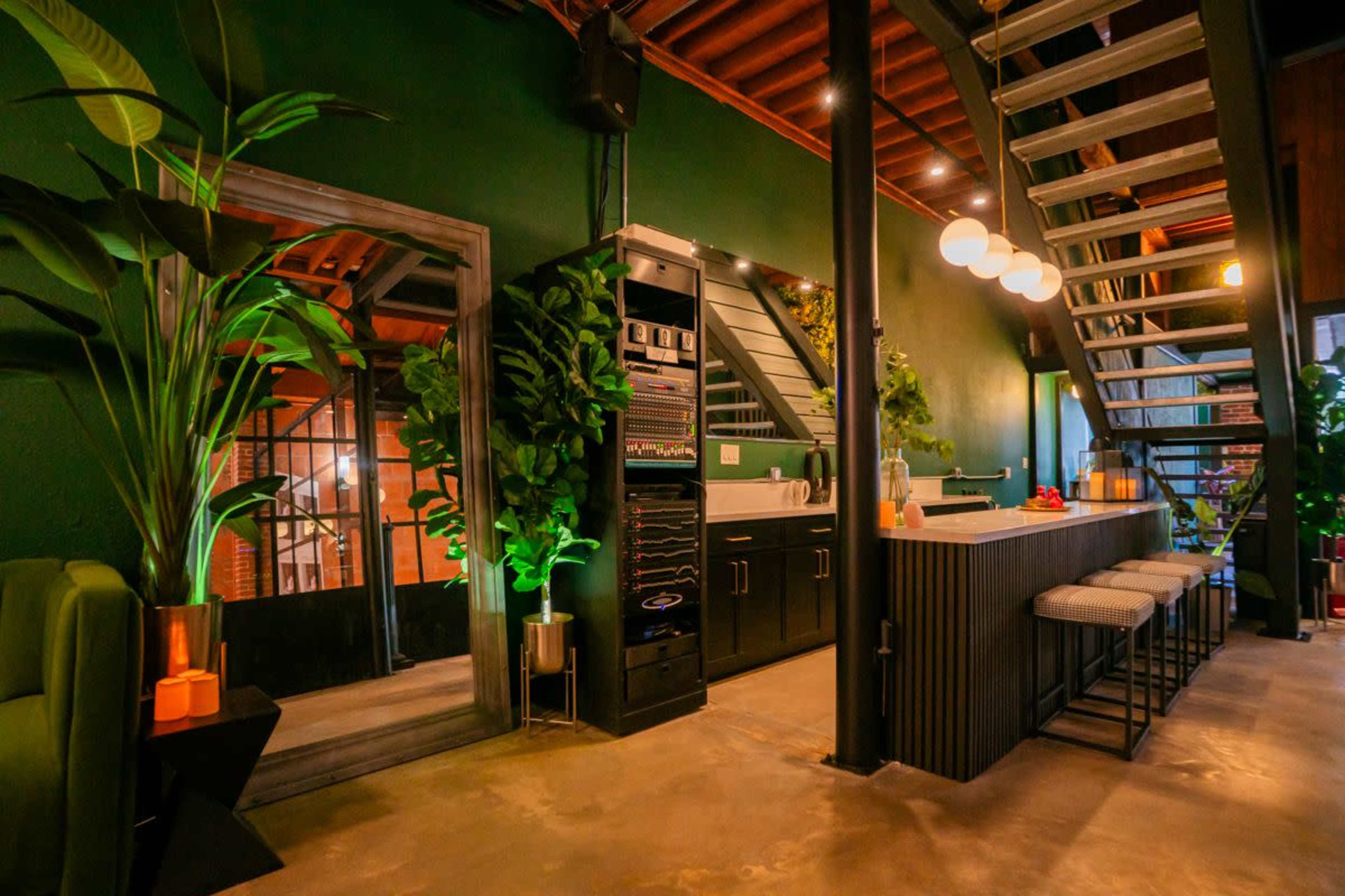 The image shows a modern, two-level interior of a café or lounge with green walls, a staircase, and a bar area featuring seating and decorative plants.