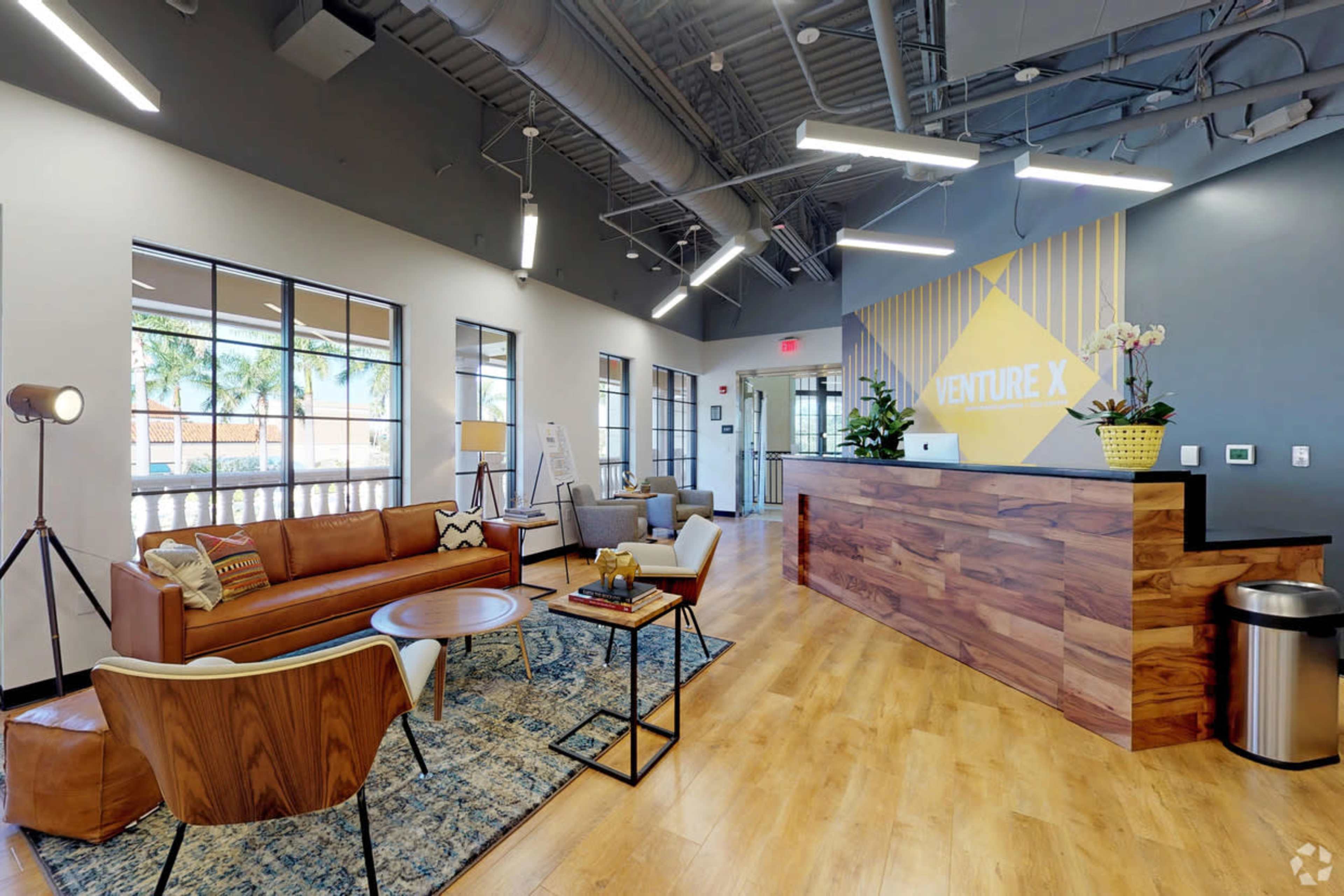 The image shows a modern office lobby with a wooden reception desk, comfortable seating areas, large windows, and decorative plants.