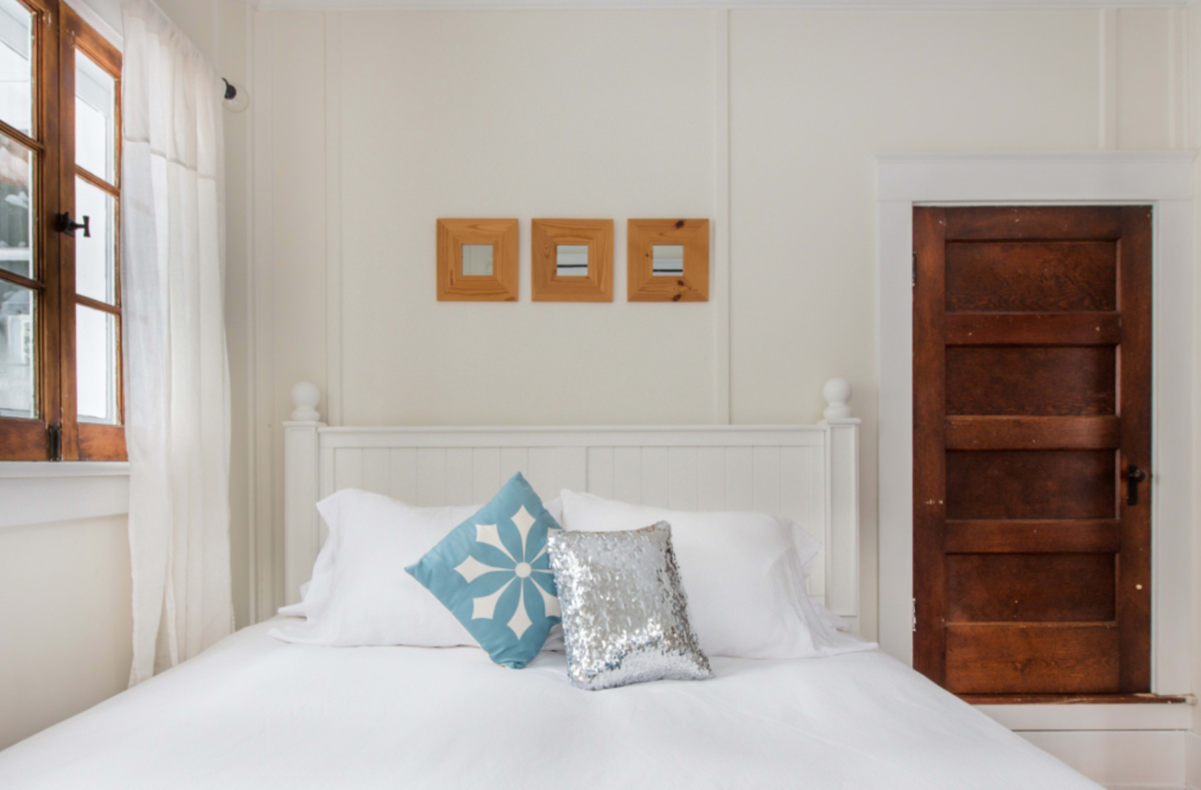 A bed with white bedding is positioned against a wall, with a decorative pillow and a sequined cushion on it, alongside a wooden door and framed mirrors on the wall.