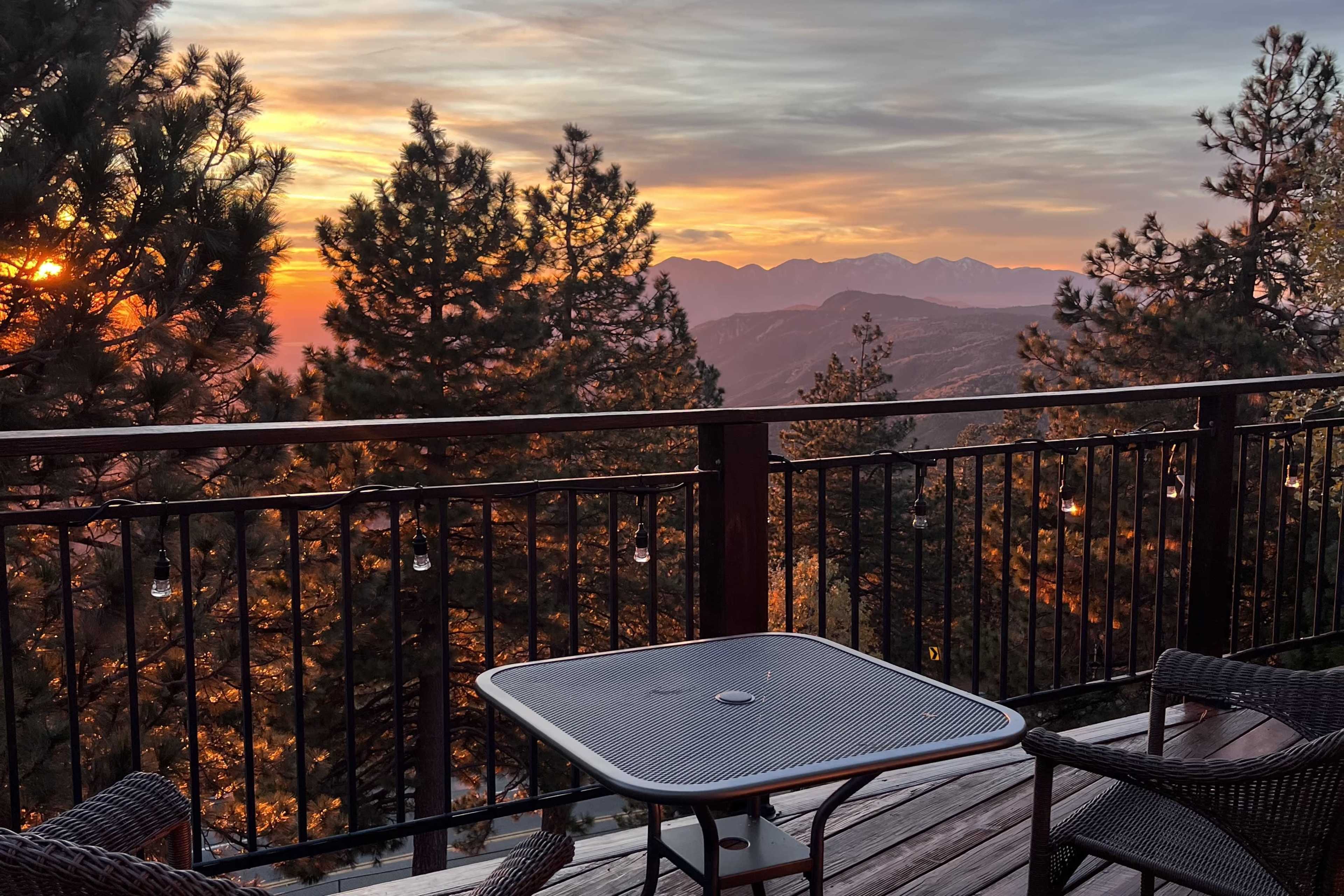 A wooden deck with a metal table and chairs overlooks a mountainous landscape at sunset, framed by pine trees.