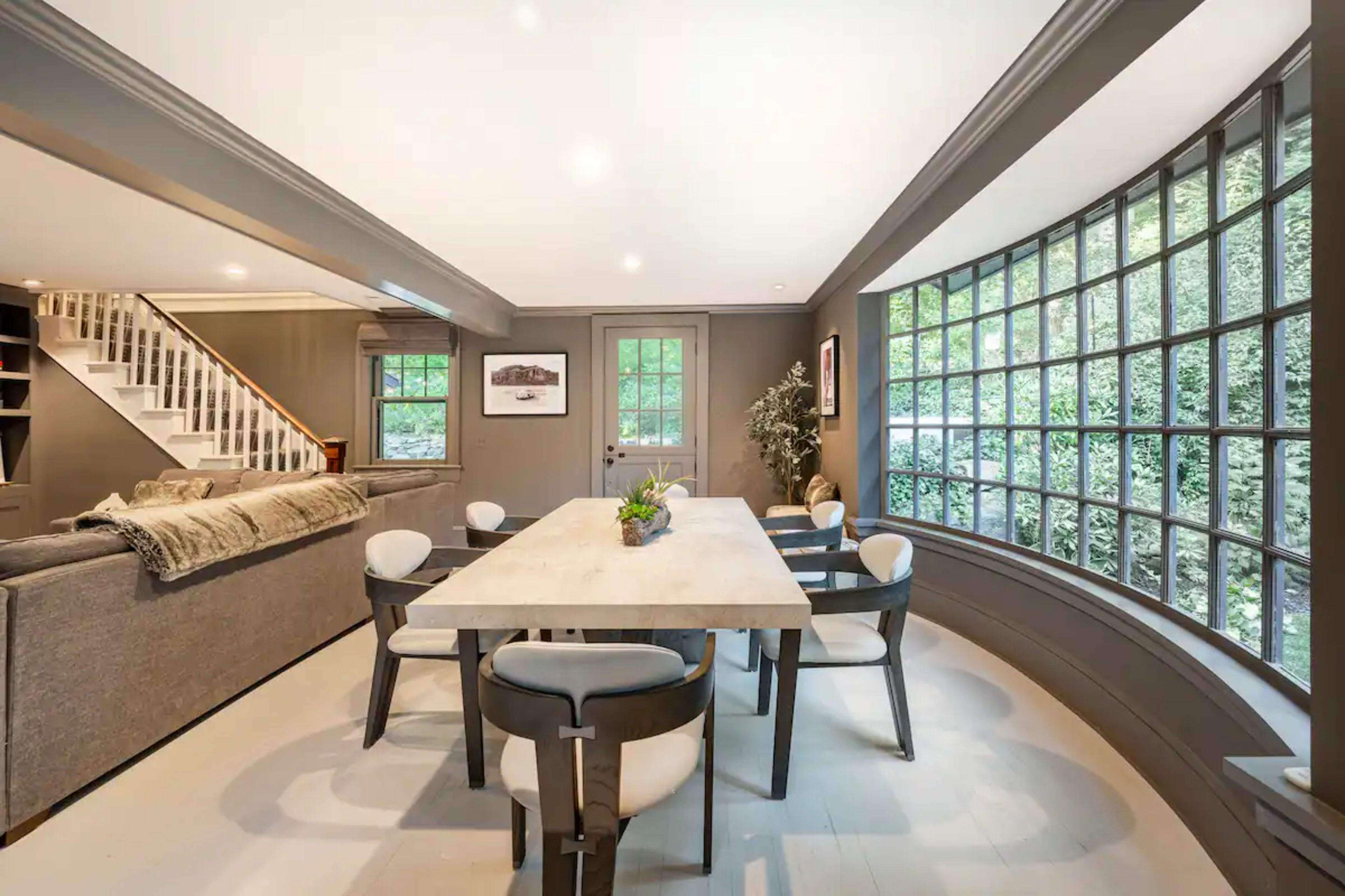 A contemporary dining area with a large table surrounded by chairs, next to a wall of windows that overlooks a green garden.