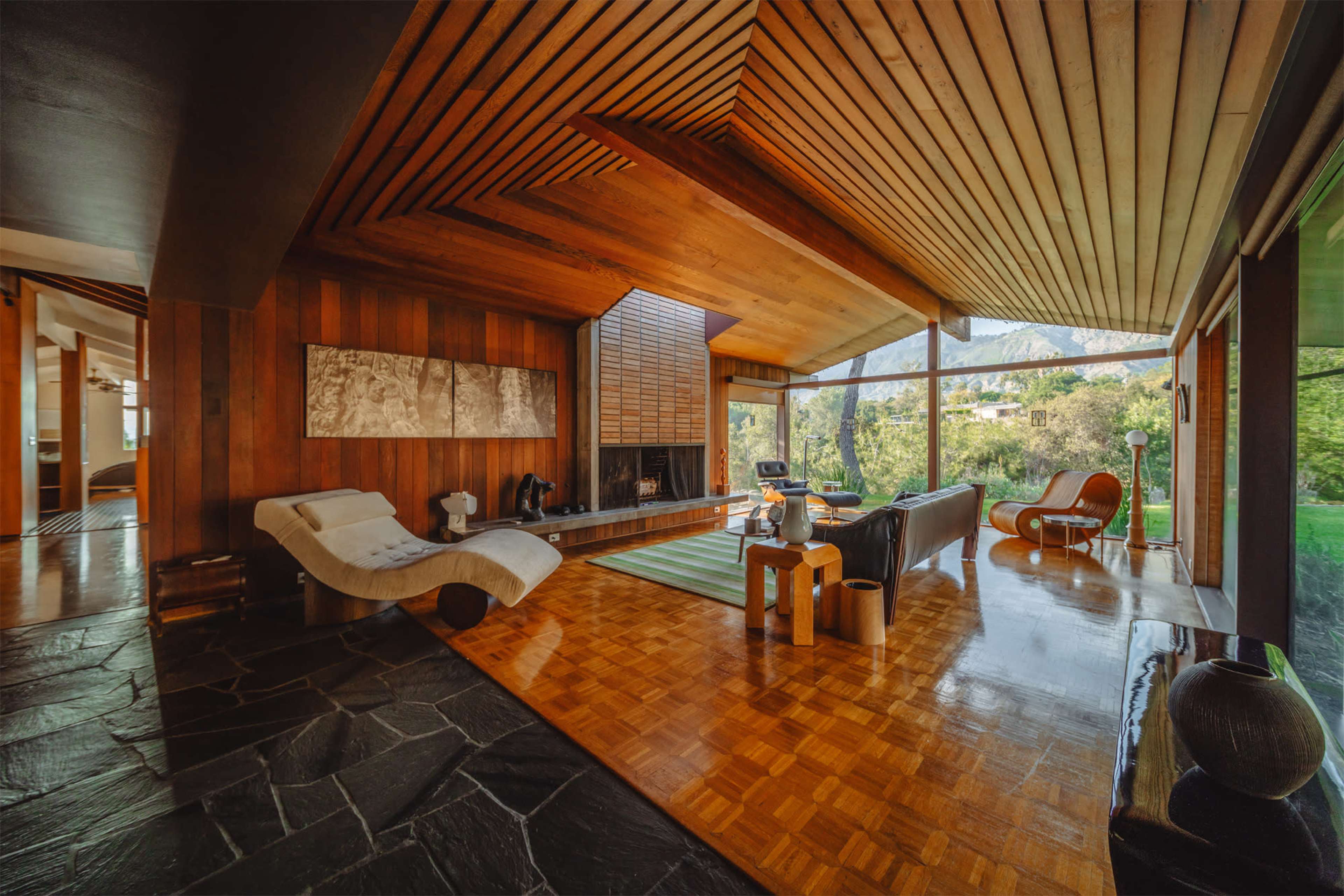 The image shows a modern living room with wooden paneling, a stone fireplace, large windows overlooking greenery, and contemporary furniture.