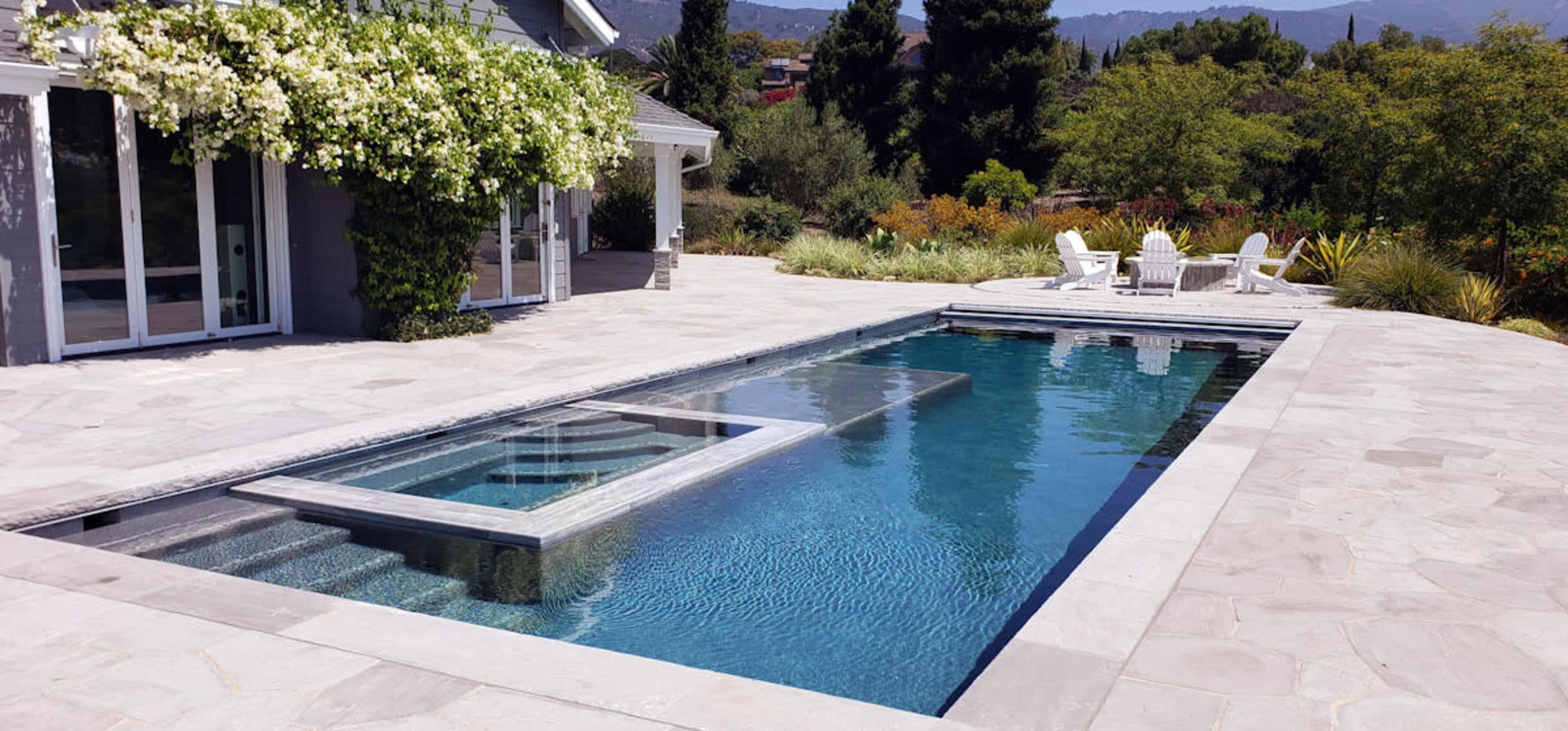 A modern stone patio with a rectangular swimming pool and a seating area featuring white chairs and a table amidst a landscaped yard.