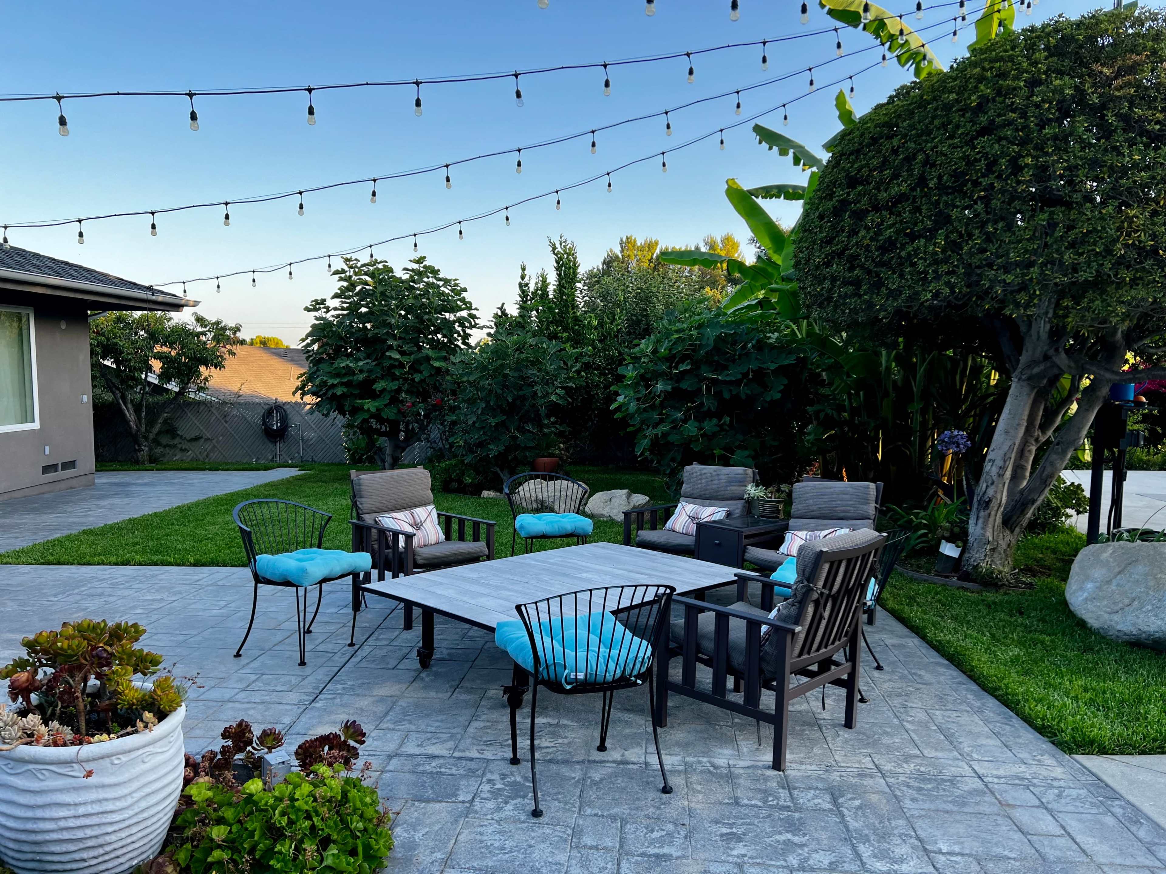 A patio area features a rectangular table surrounded by six chairs with blue cushions, illuminated by string lights overhead.