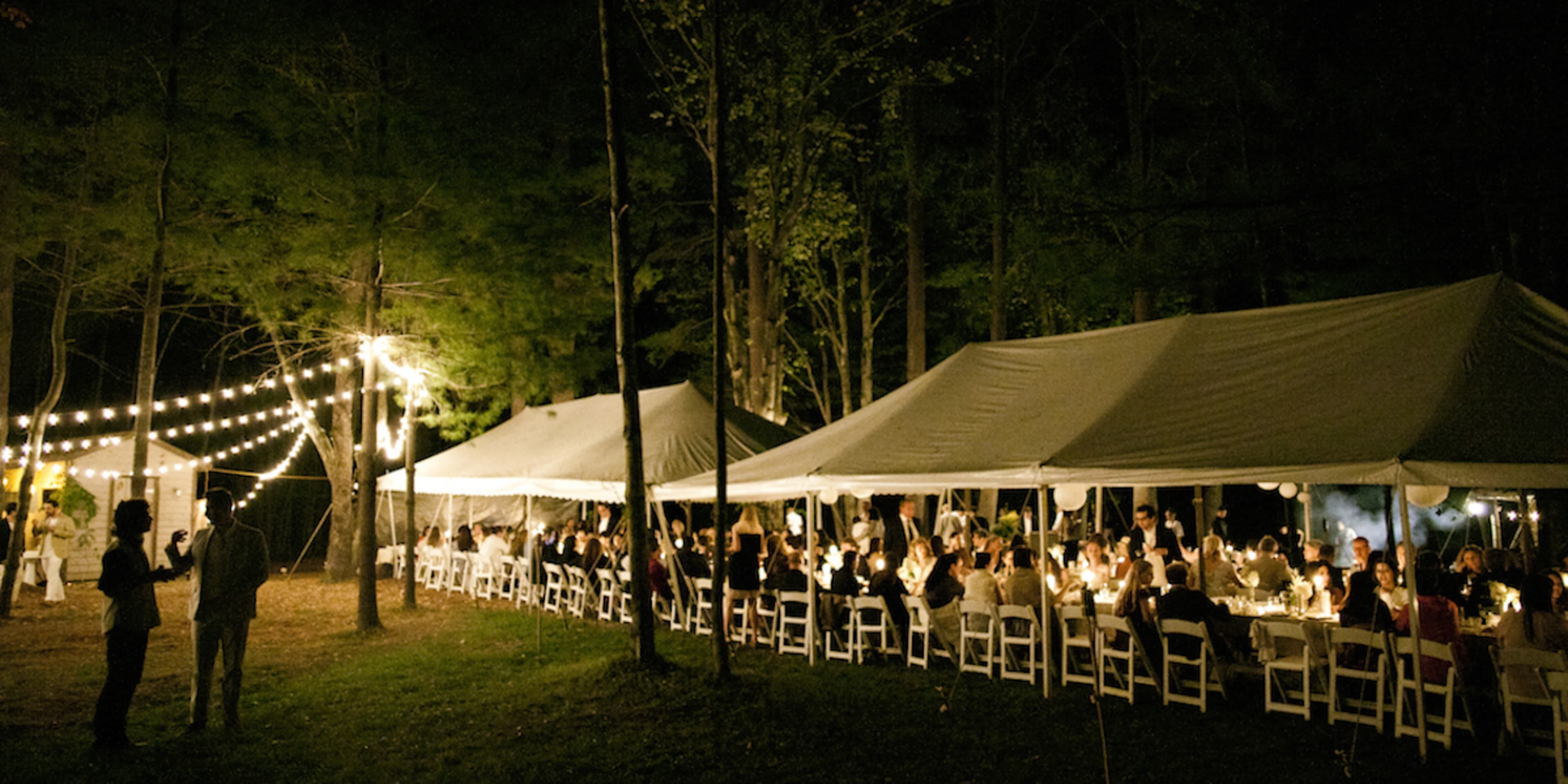 A group of people gather under string lights at tables arranged beneath large tents in a forested setting at night.