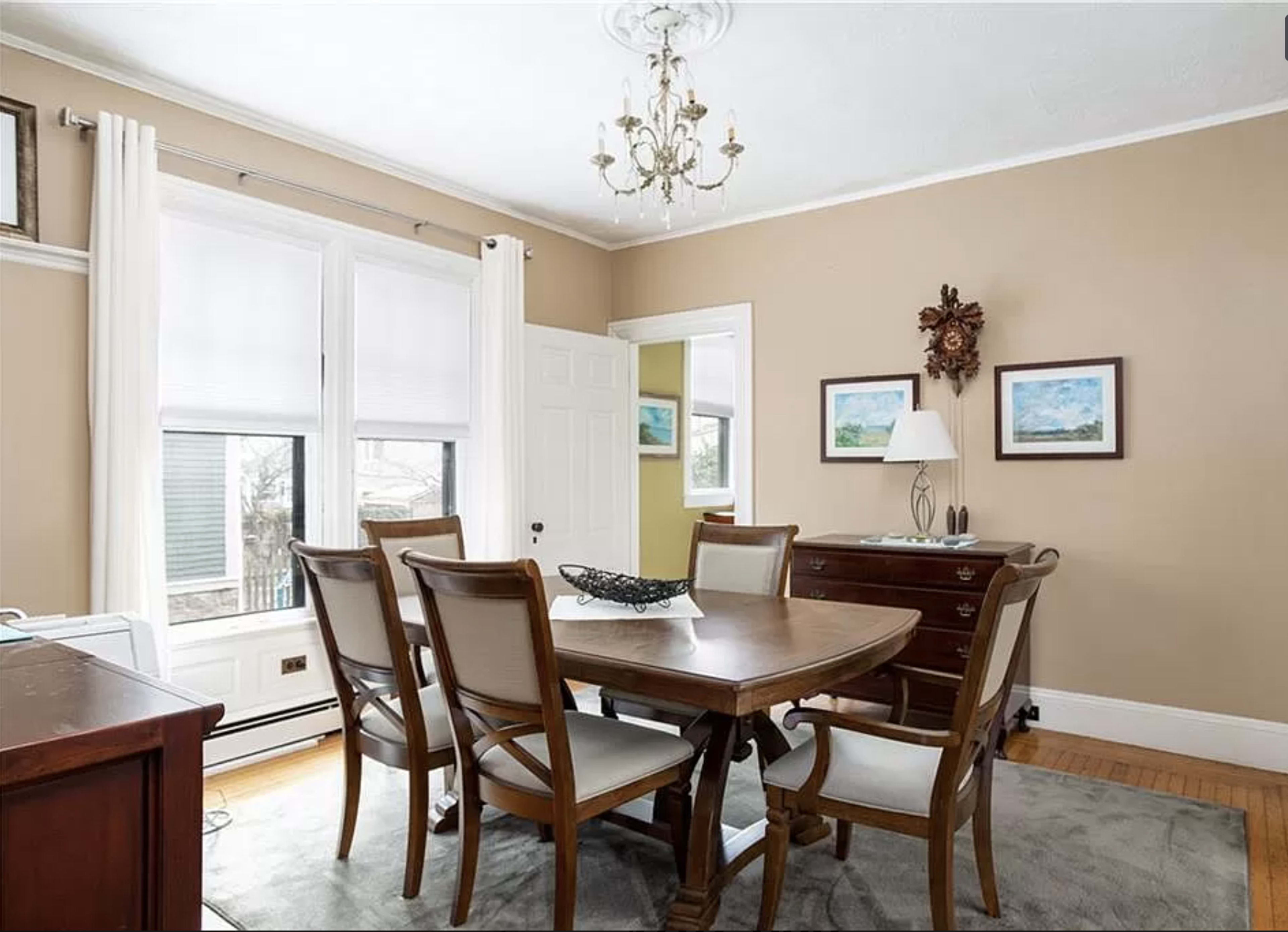 The image shows a dining room featuring a wooden table surrounded by six chairs, a sideboard, and a chandelier hanging from the ceiling.