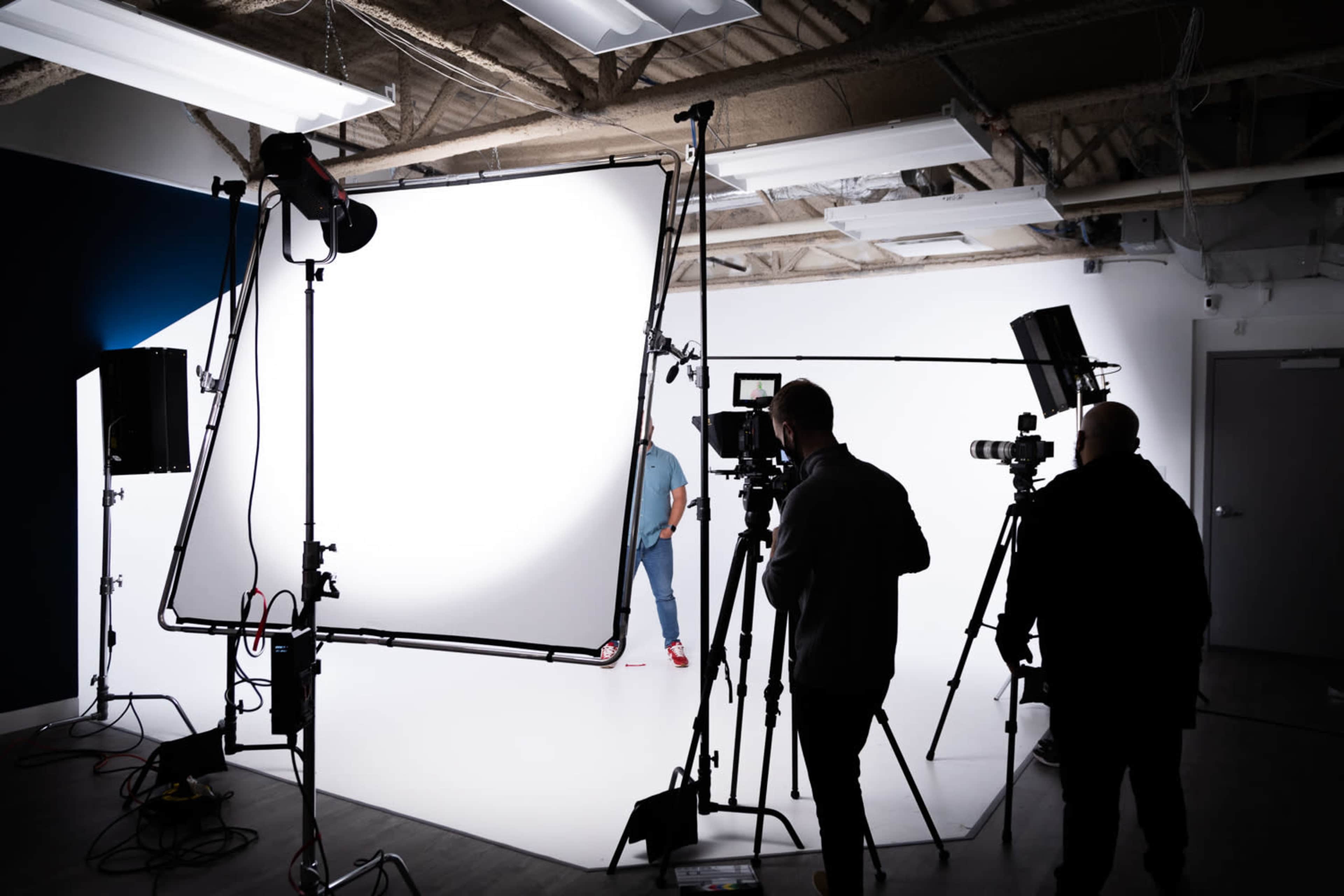 A photography studio is set up with multiple light sources and camera equipment focused on a person in front of a white backdrop.
