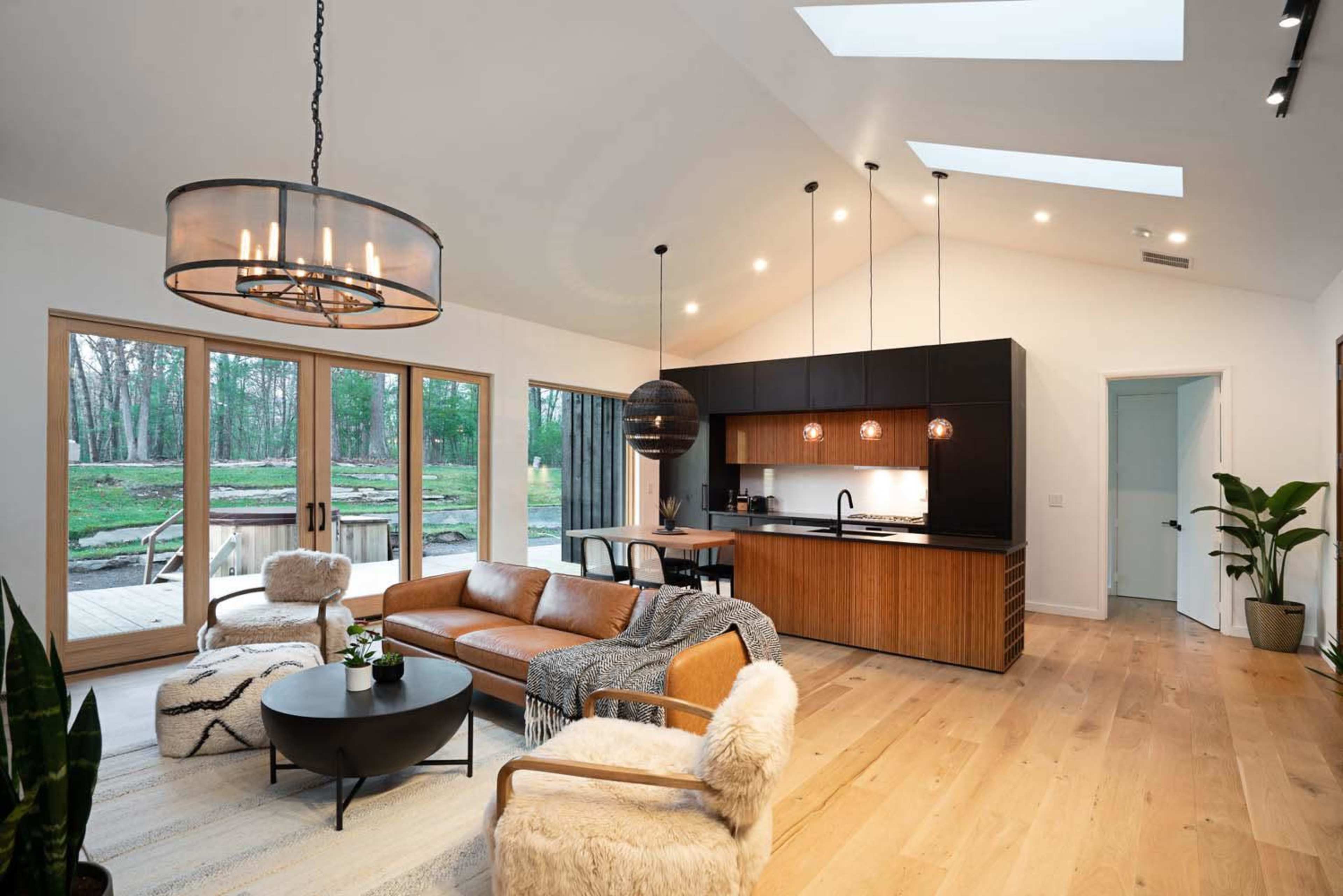 A modern living room with a leather sofa, a round coffee table, and a kitchen area featuring dark cabinetry and large windows that open to a wooded outdoor space.
