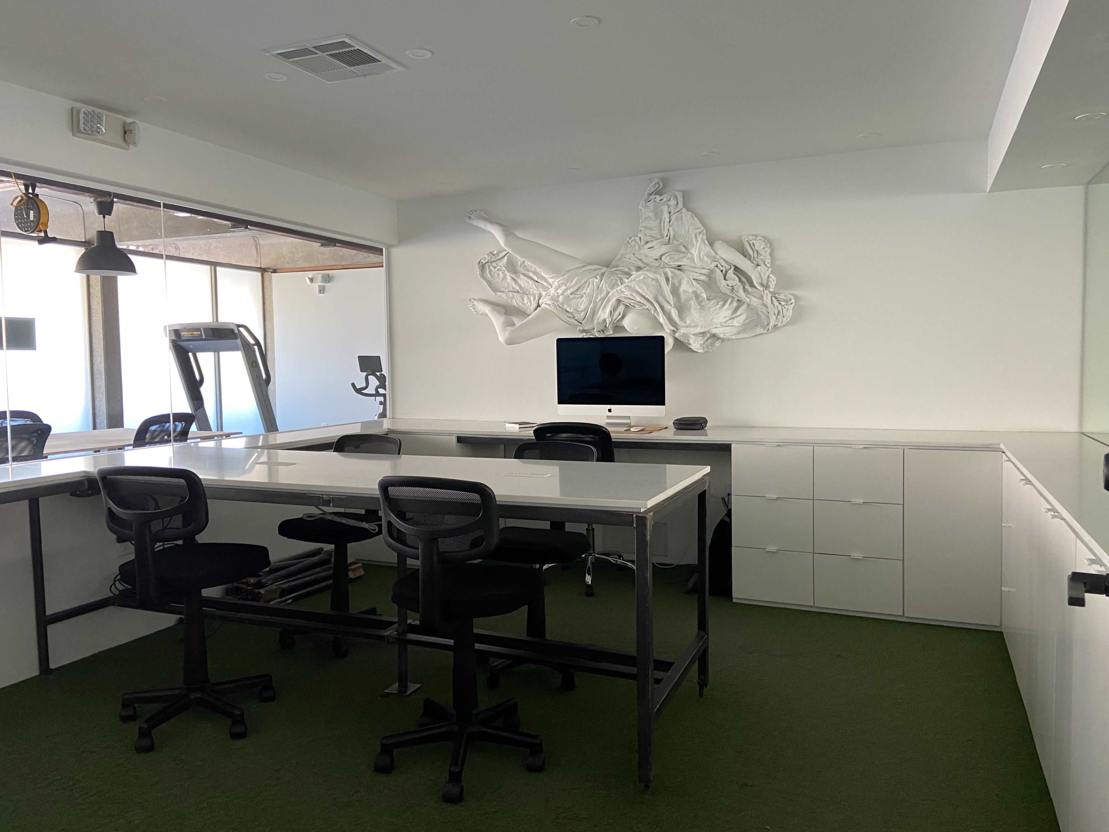 A modern office space features a large white wall sculpture, a desk with chairs, and a treadmill in the background.