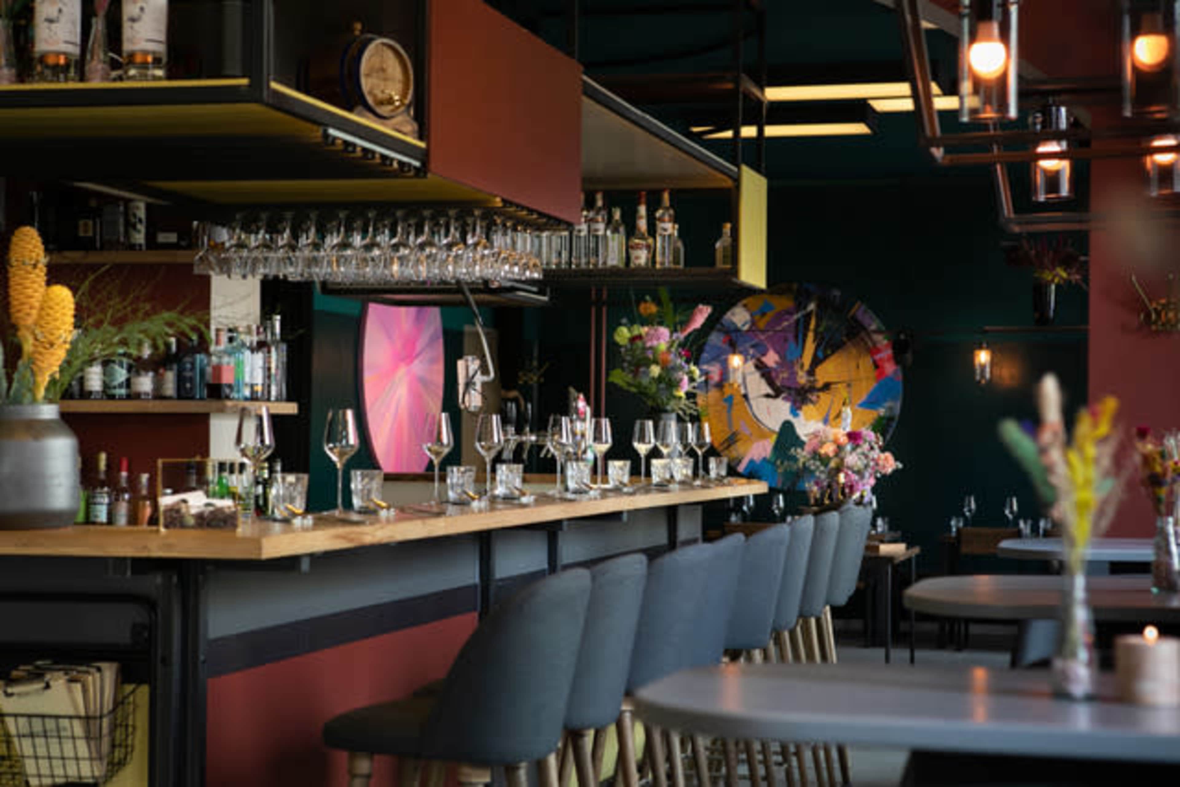 The image shows a modern bar area with a long counter, glassware neatly arranged, and colorful decor featuring plants and artistic elements.