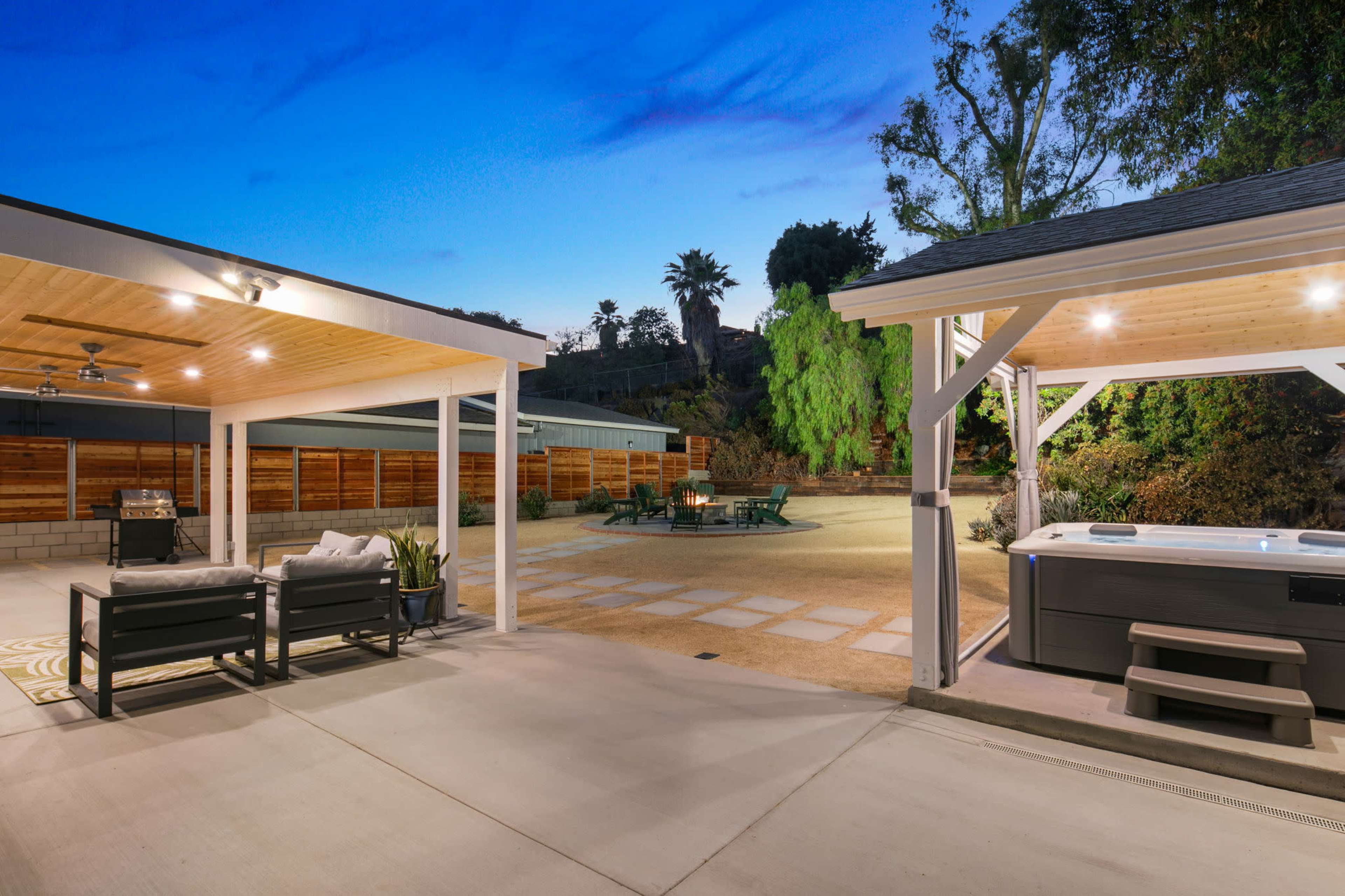The scene shows a spacious outdoor area featuring a covered patio with seating, a fire pit surrounded by benches, and a hot tub, all set against a backdrop of trees and a blue evening sky.