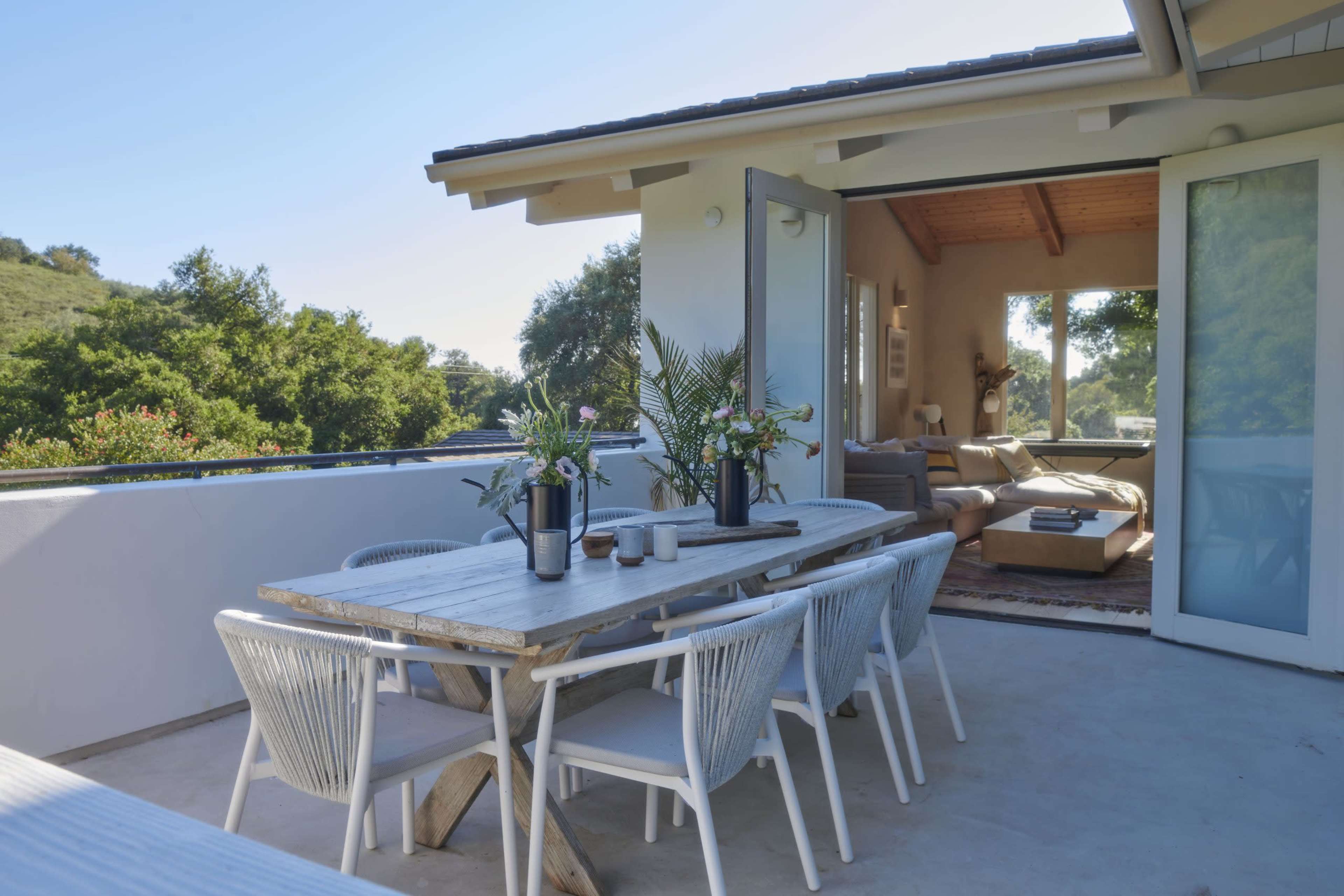 A spacious outdoor dining area features a long wooden table with chairs, surrounded by greenery and connected to a modern living space.