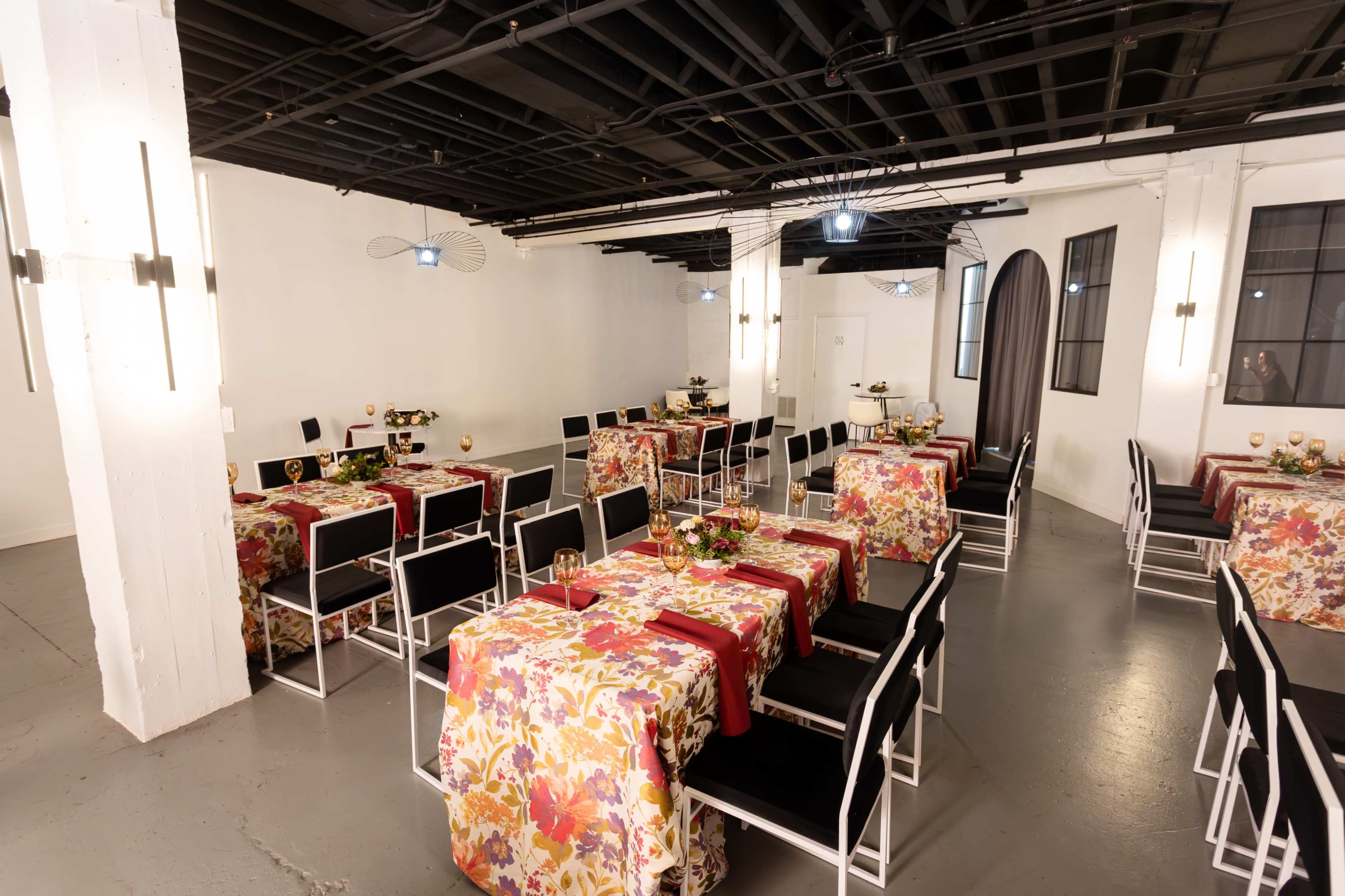 The image shows a modern event space arranged with tables covered in floral tablecloths and surrounded by black chairs.