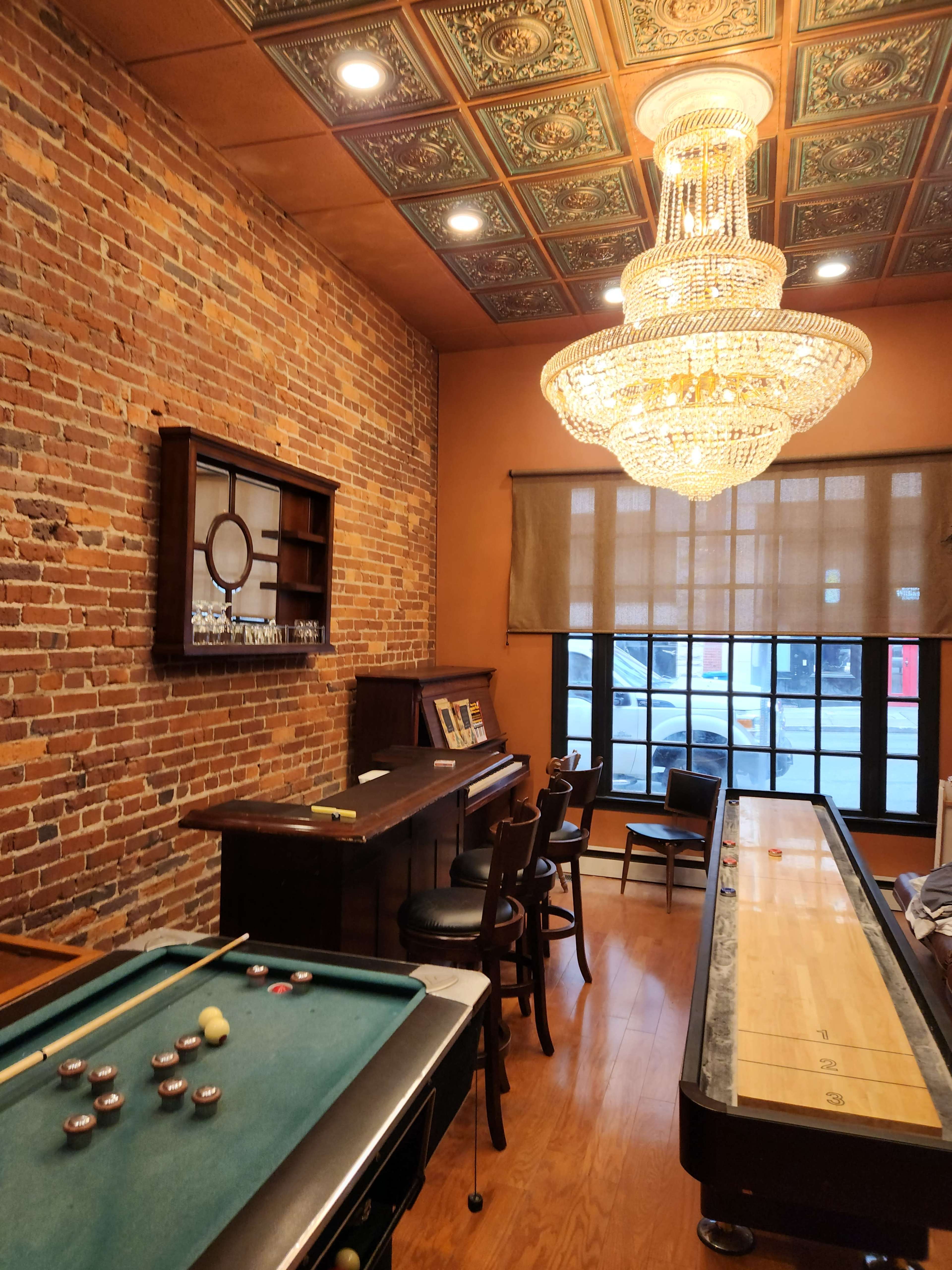 The image shows a game room featuring a pool table, a shuffleboard table, a wooden bar with chairs, and a large chandelier hanging from a decorative ceiling.