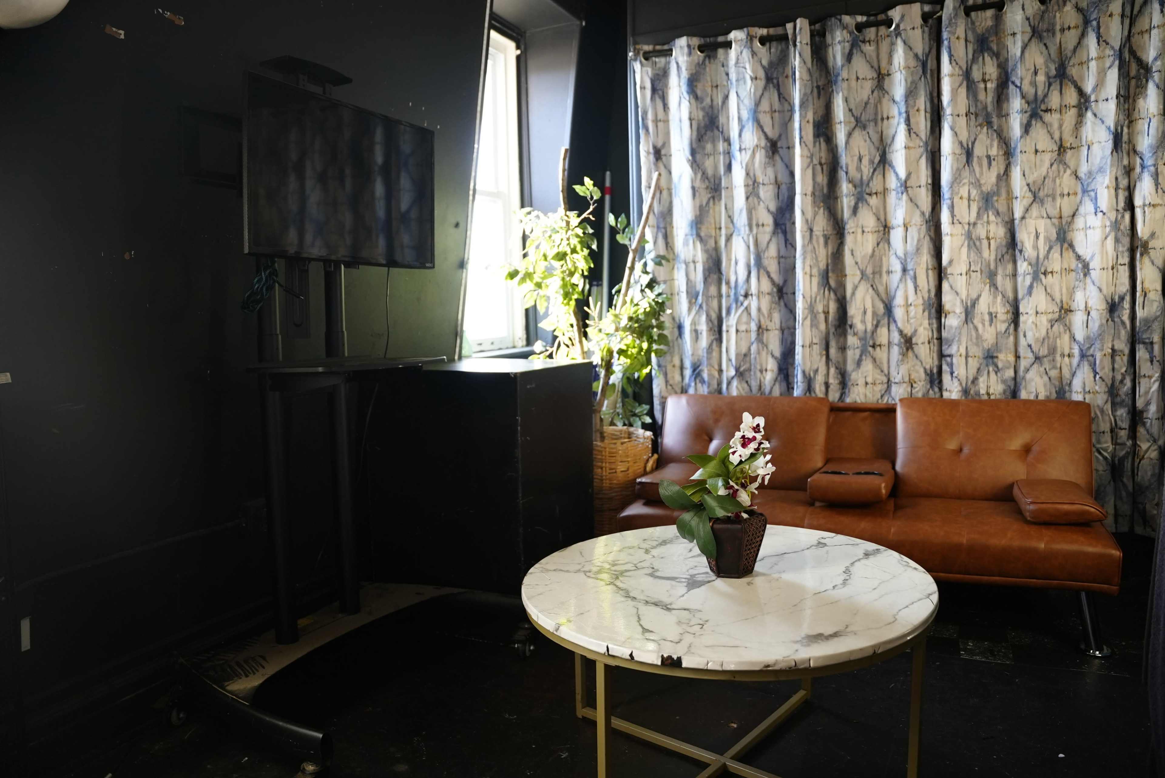 The image shows a small lounge area with a brown leather couch, a marble-top coffee table, a potted plant, and a TV mounted on a stand beside a window draped with a patterned curtain.