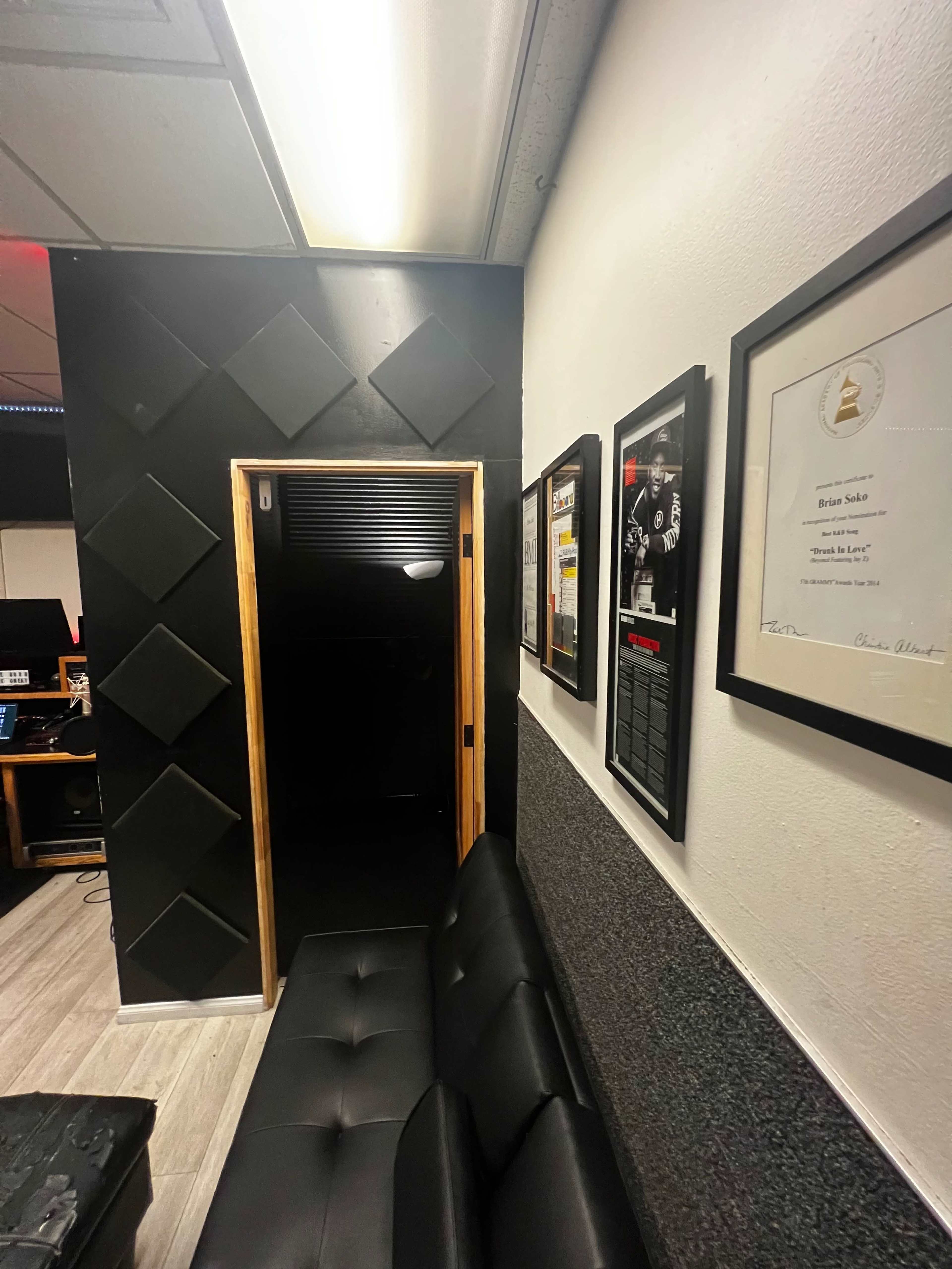 The image shows a dimly lit hallway with a wooden door leading to another room, flanked by framed awards and a black leather couch against a textured wall.