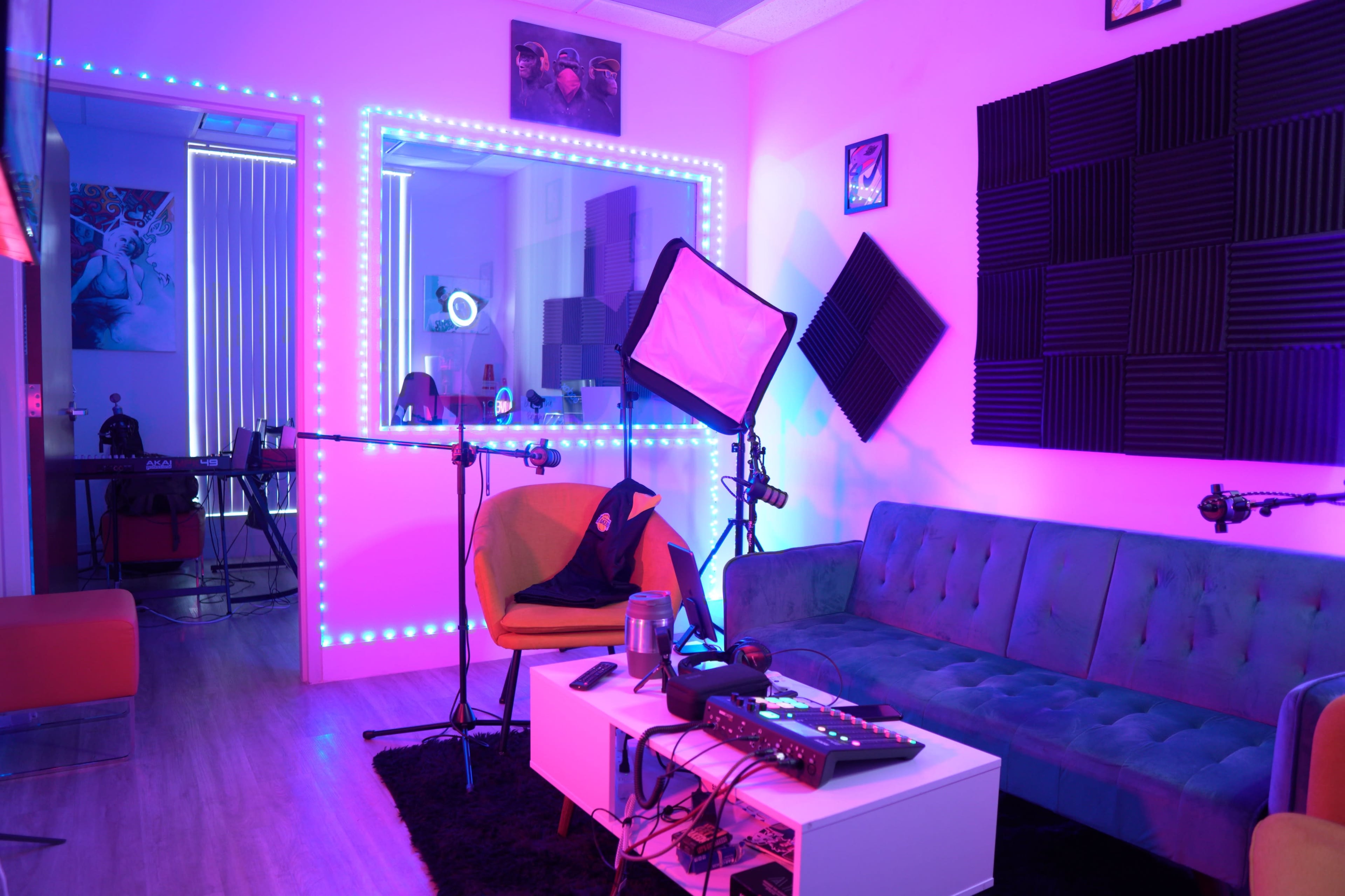 A brightly lit recording studio with neon lights, a green couch, a coffee table with equipment, and soundproofing panels on the walls.