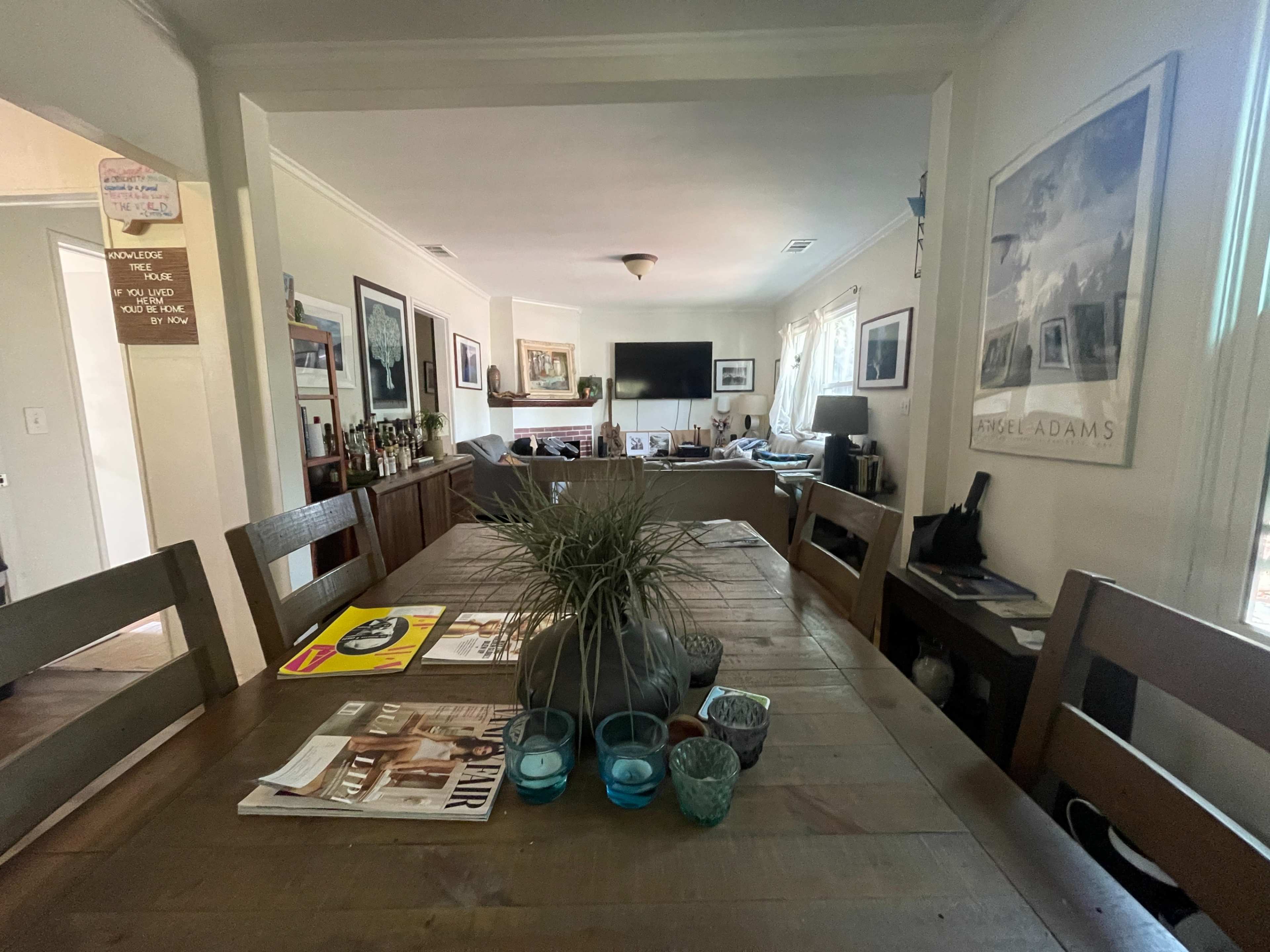 A dining area with a wooden table, decorative items, and a view into a living room with a television and various artworks on the walls.