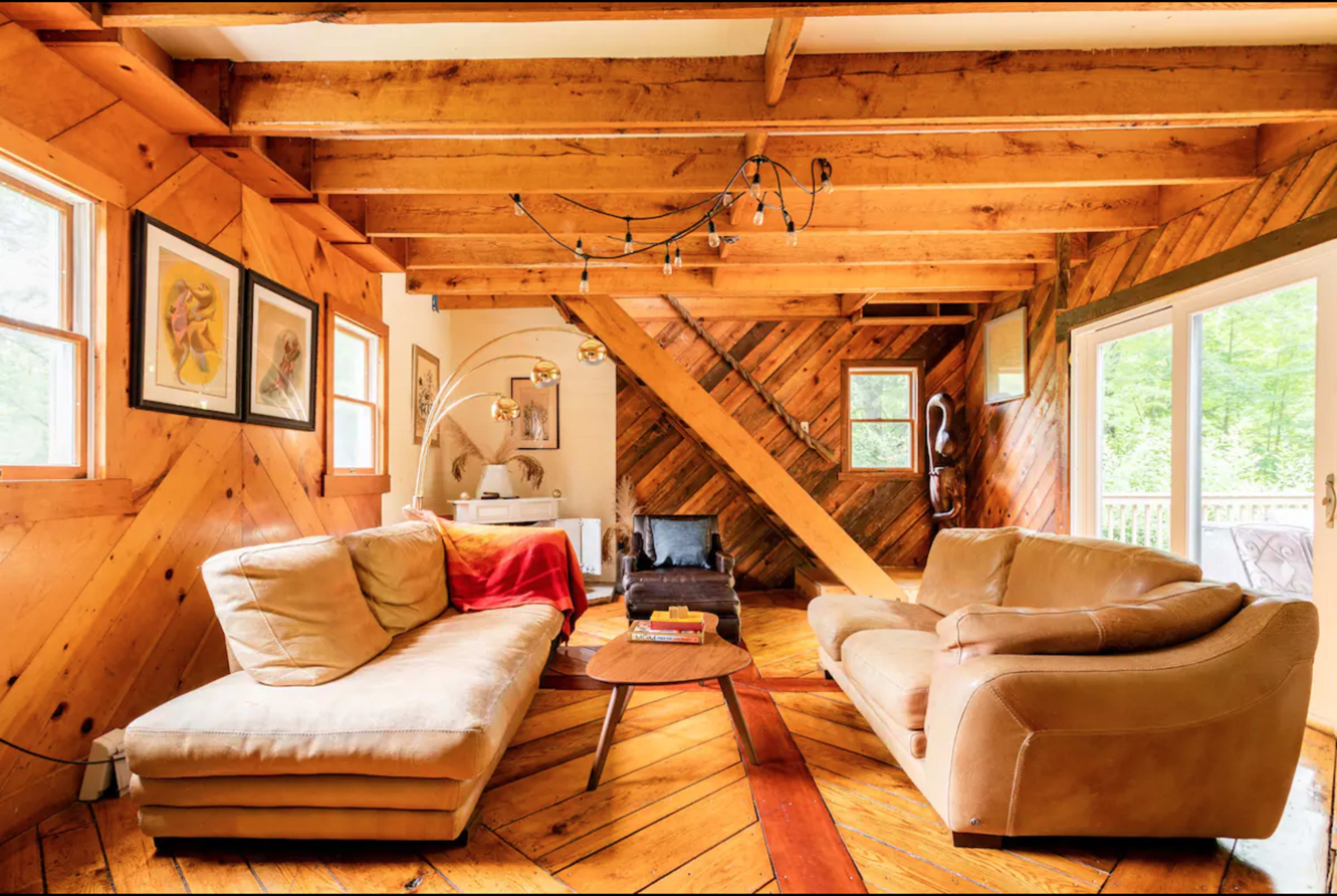 The image shows a cozy living room with wooden beams and diagonal wood paneling, featuring two sofas, a circular coffee table, and a staircase.