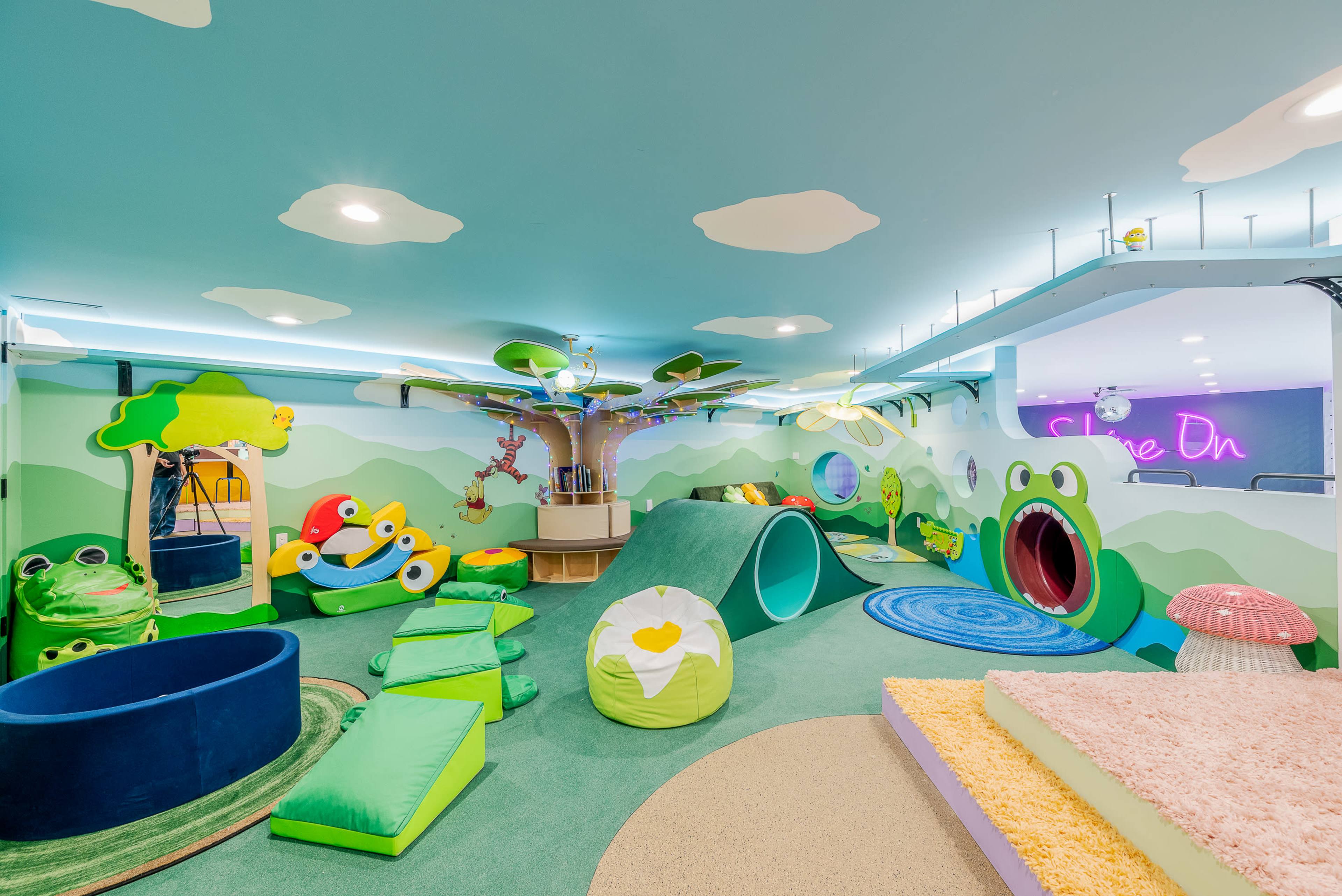 The image depicts a brightly colored children's playroom featuring various plush, frog-themed structures and a large tree centerpiece, with soft seating and tunnels for play.
