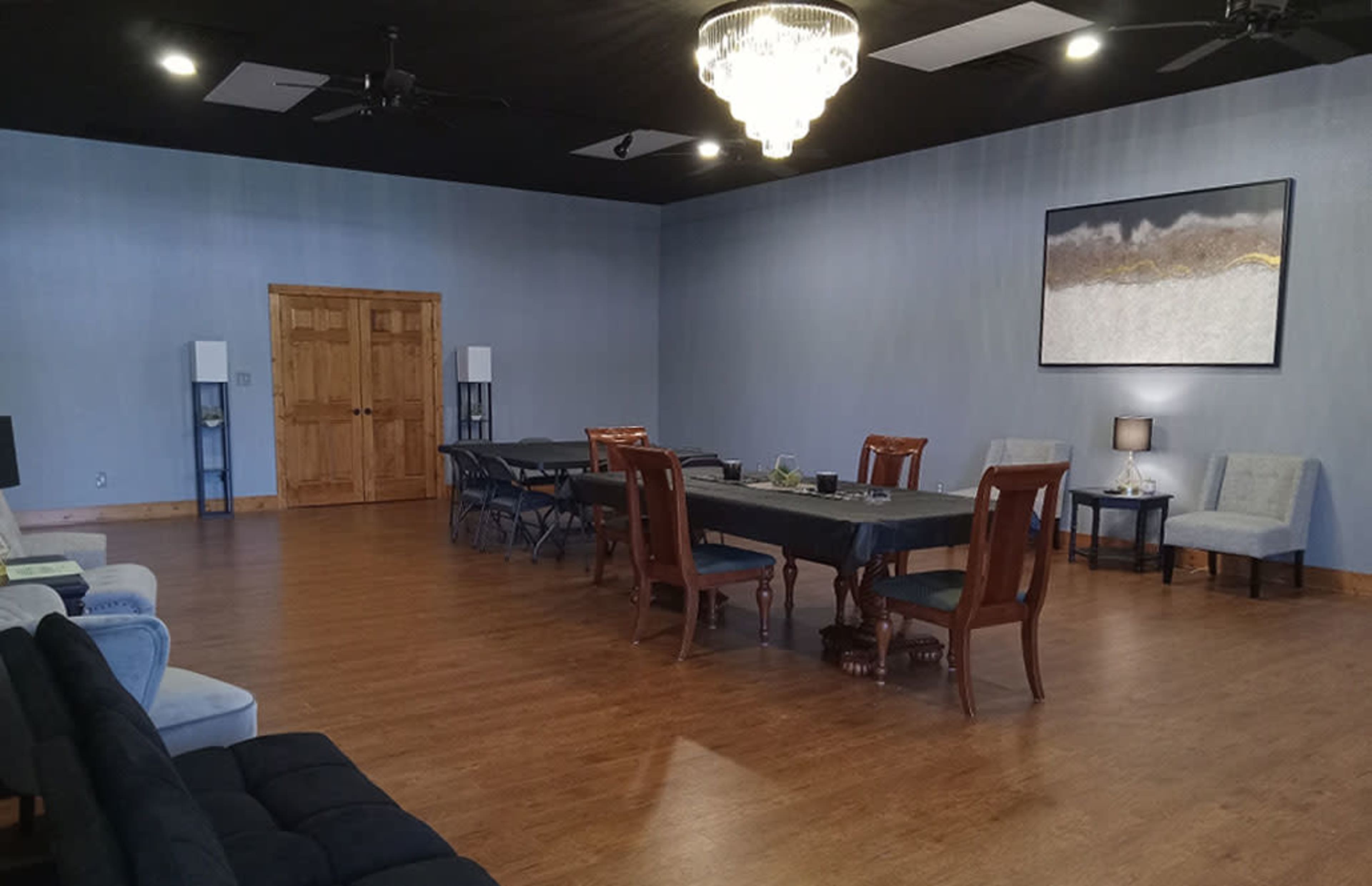 The image shows a spacious room with wooden flooring, featuring a long table surrounded by chairs, a decorative chandelier, and a few seating areas.