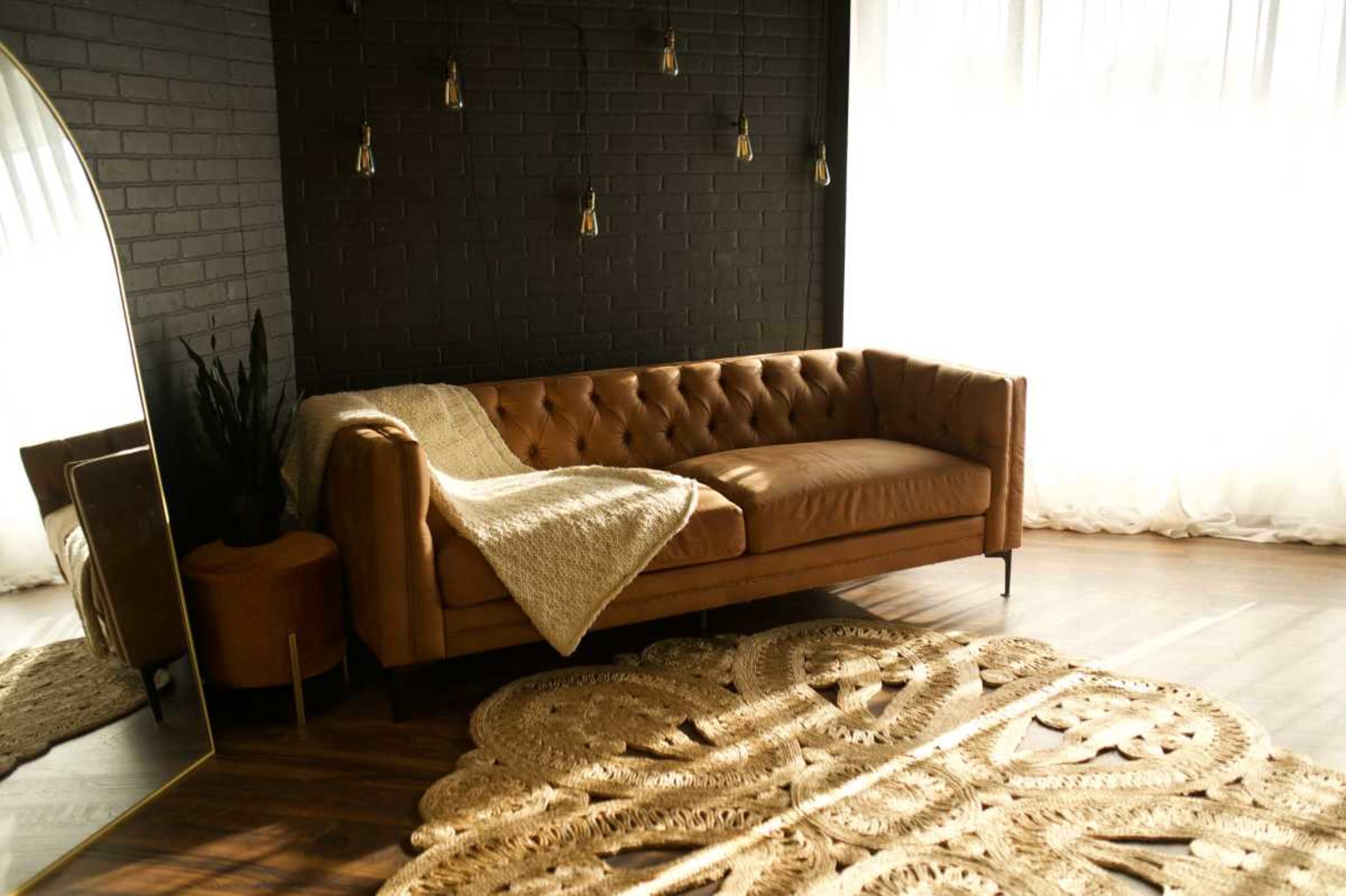 The image shows a cozy living room featuring a tan leather sofa, a large round woven rug, and natural light streaming through sheer curtains.