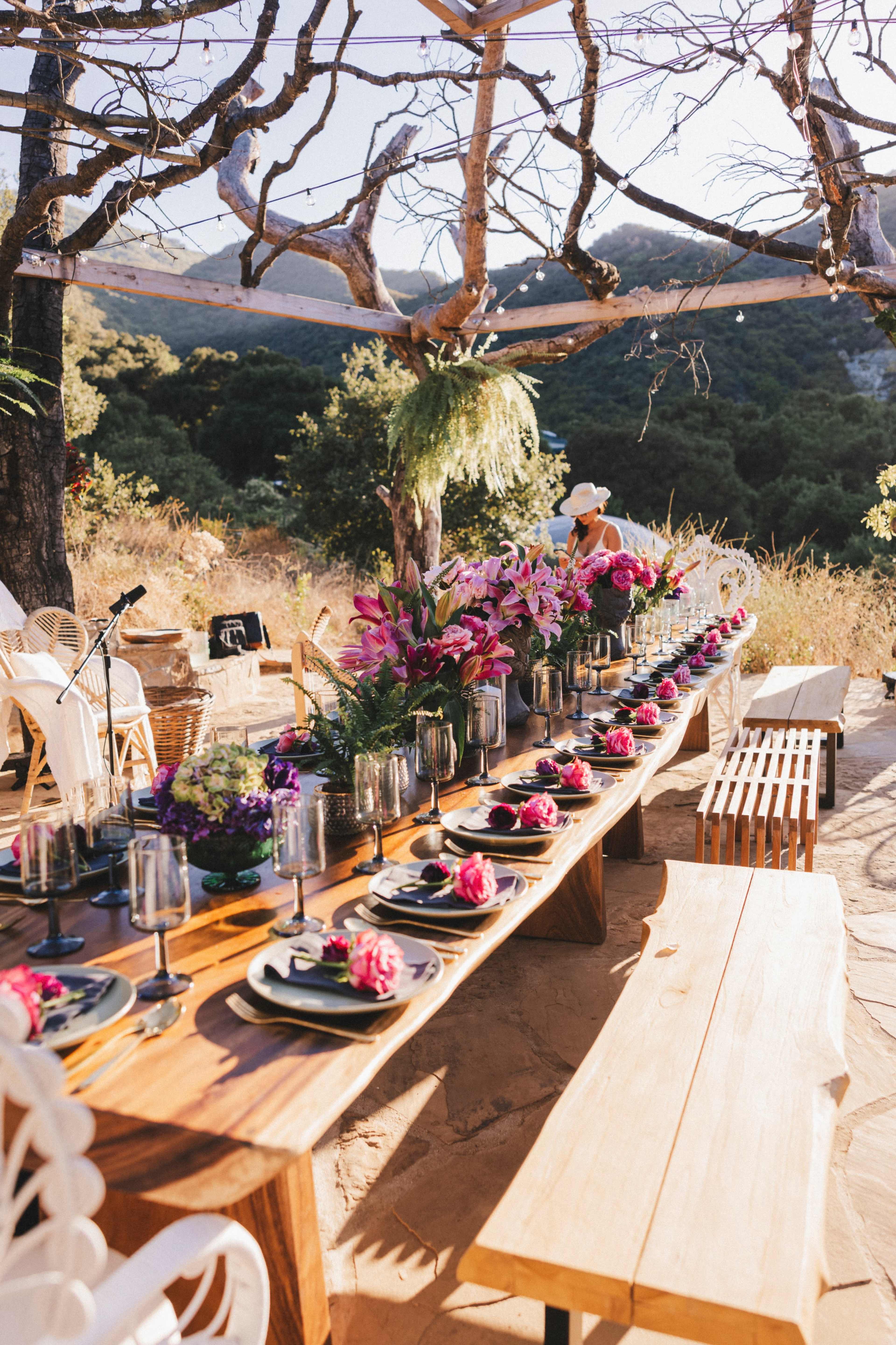 A long wooden table is set for a meal outdoors, adorned with flowers and plates, beneath a canopy of branches.