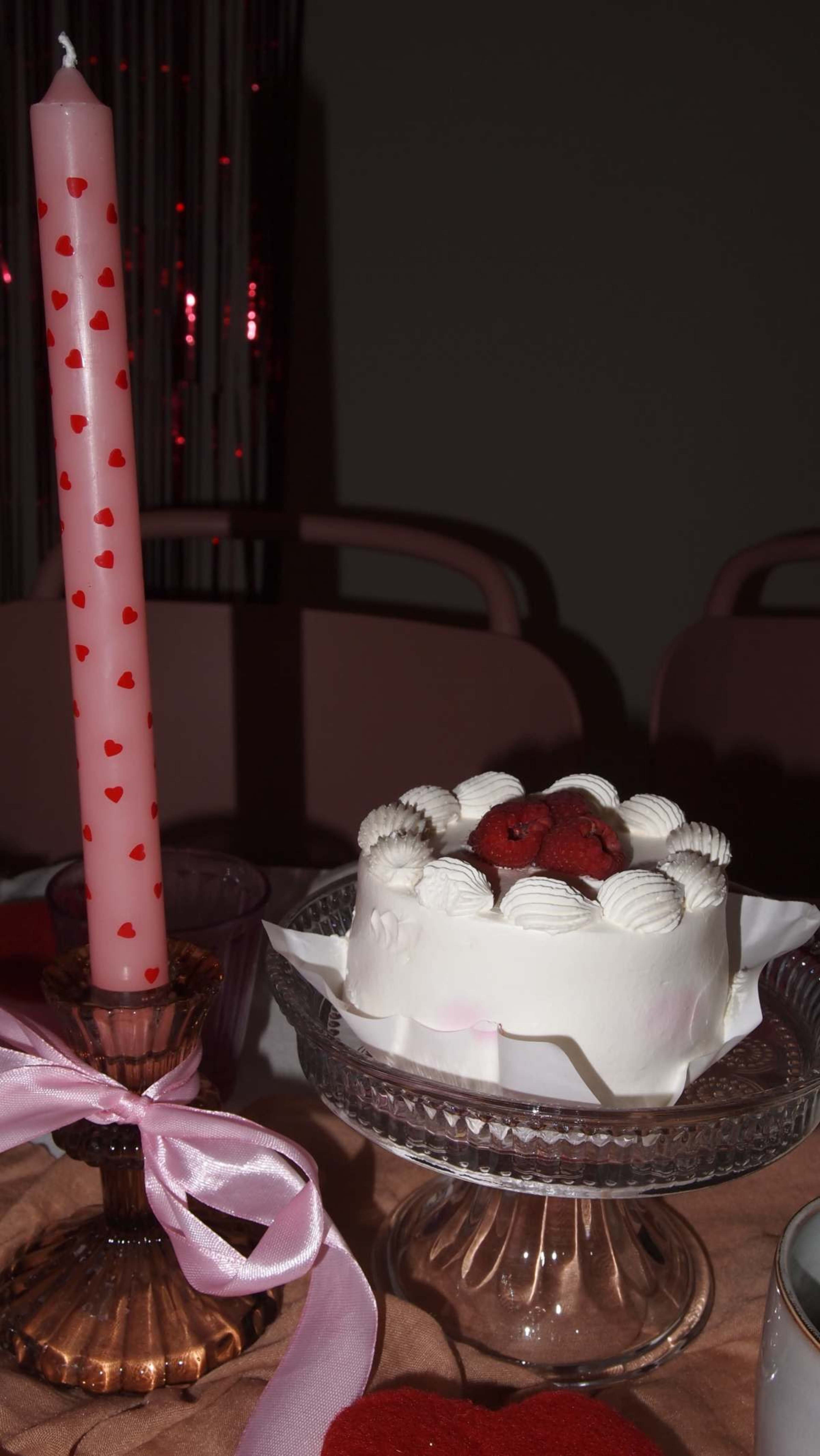 A white frosted cake with strawberries on top is displayed beside a tall pink candle adorned with red hearts.