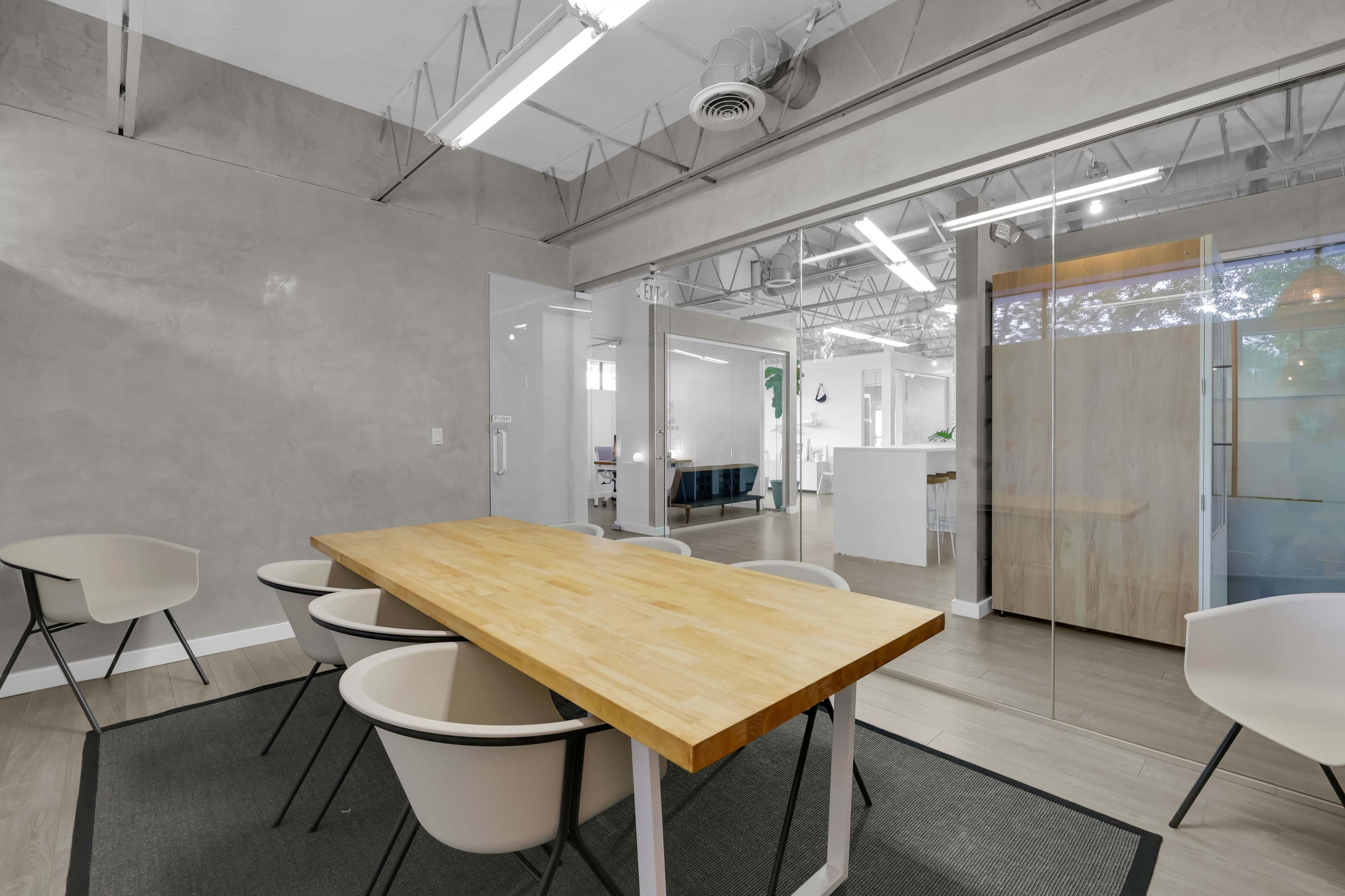 A modern office space features a wooden conference table surrounded by four light-colored chairs, with glass walls and a minimalist aesthetic.