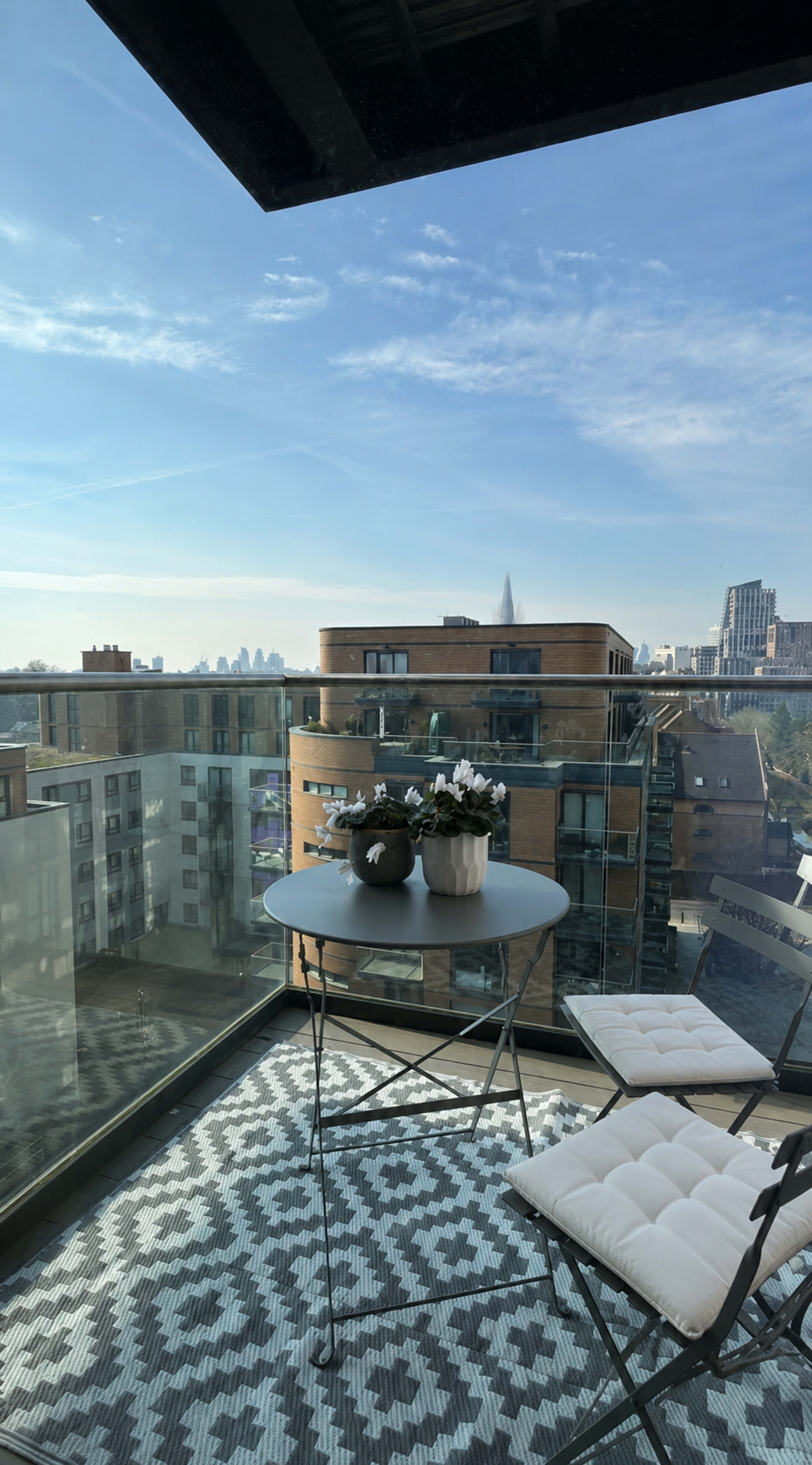 A balcony features a circular table with potted plants and two white chairs, overlooking a cityscape under a clear sky.