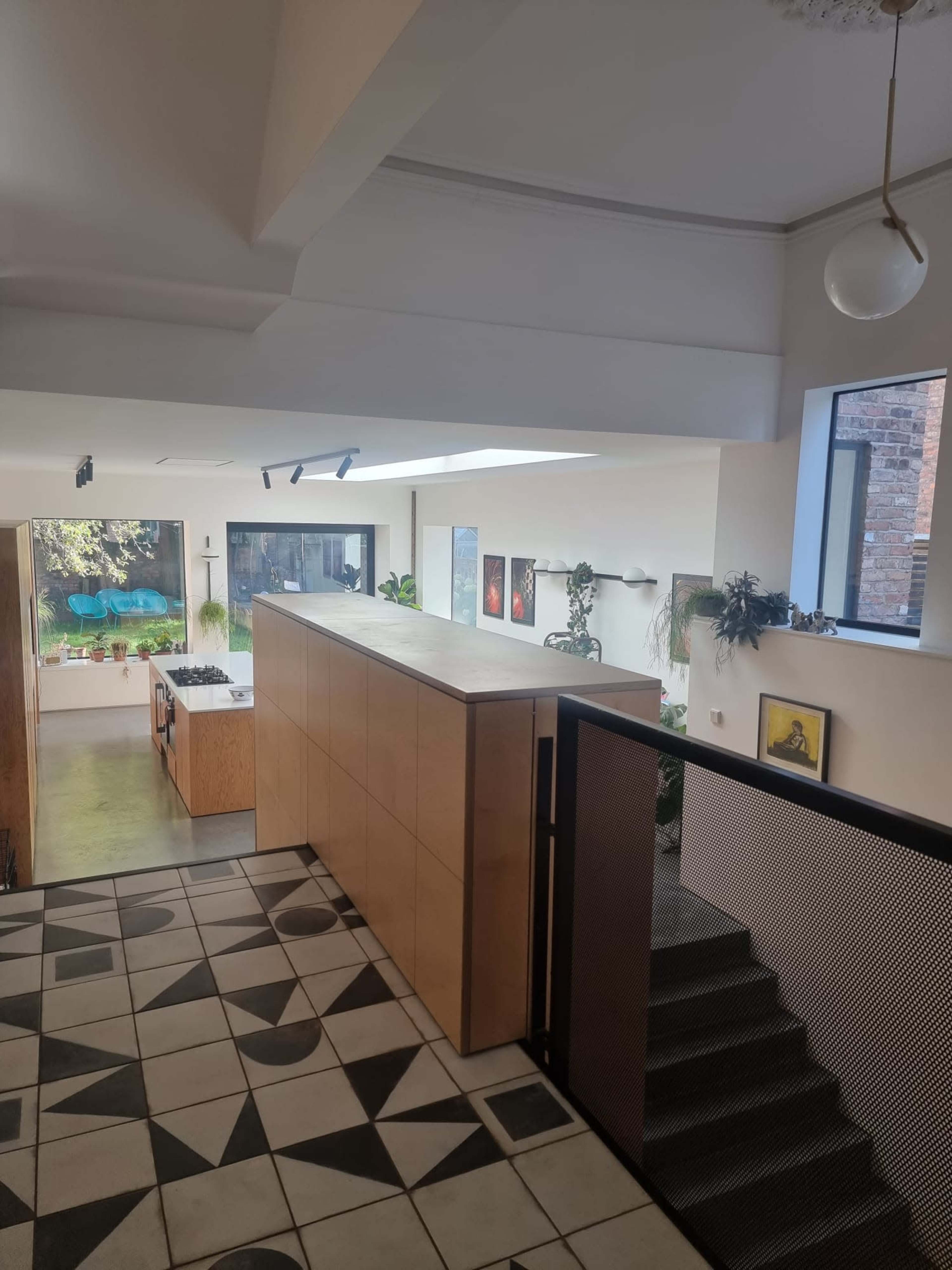 A modern interior of a home featuring a wooden kitchen island, large windows with greenery outside, and a staircase with a metal railing.