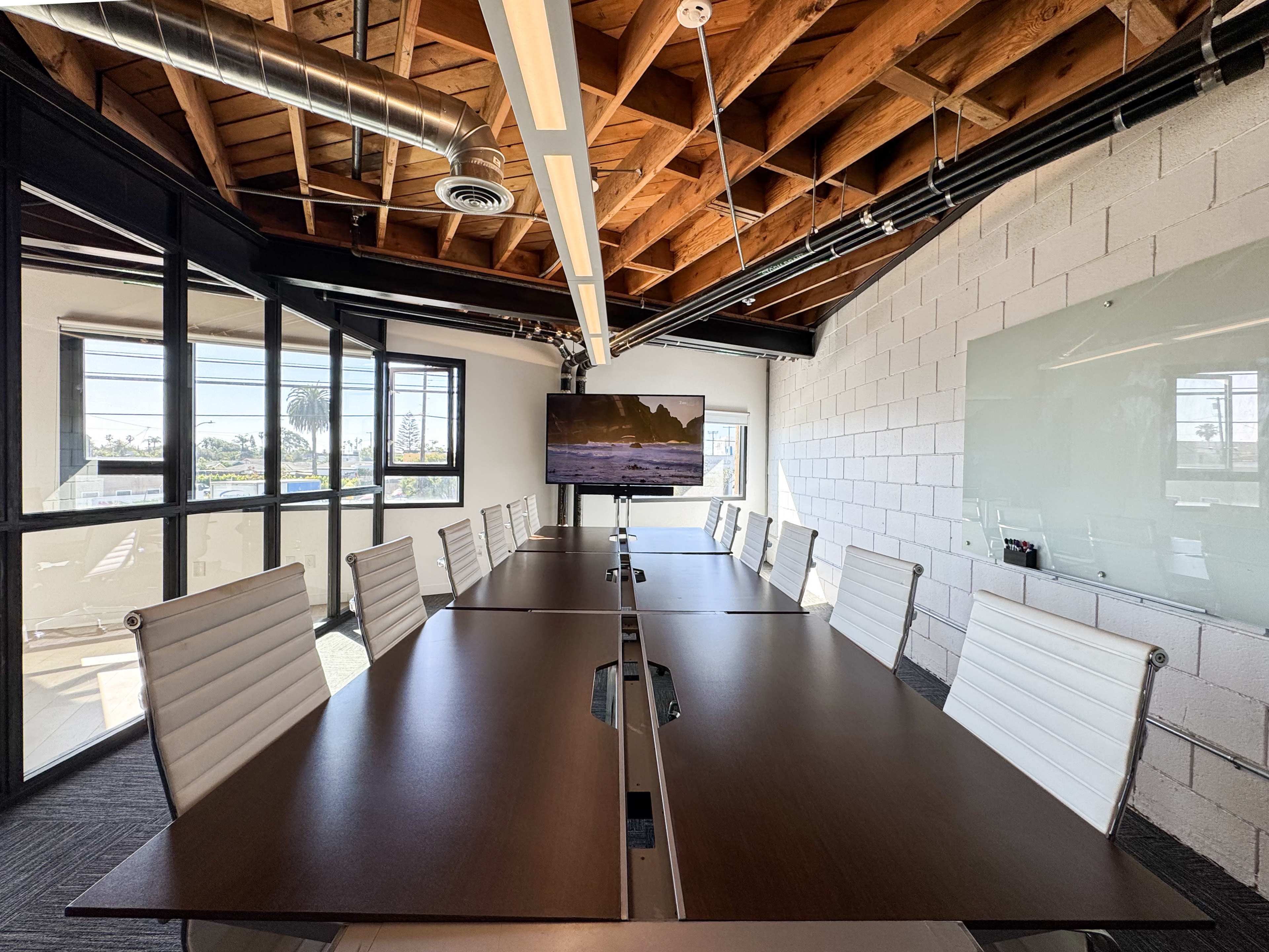 A modern conference room features a long wooden table with white chairs and a large screen at one end, illuminated by natural light from windows.