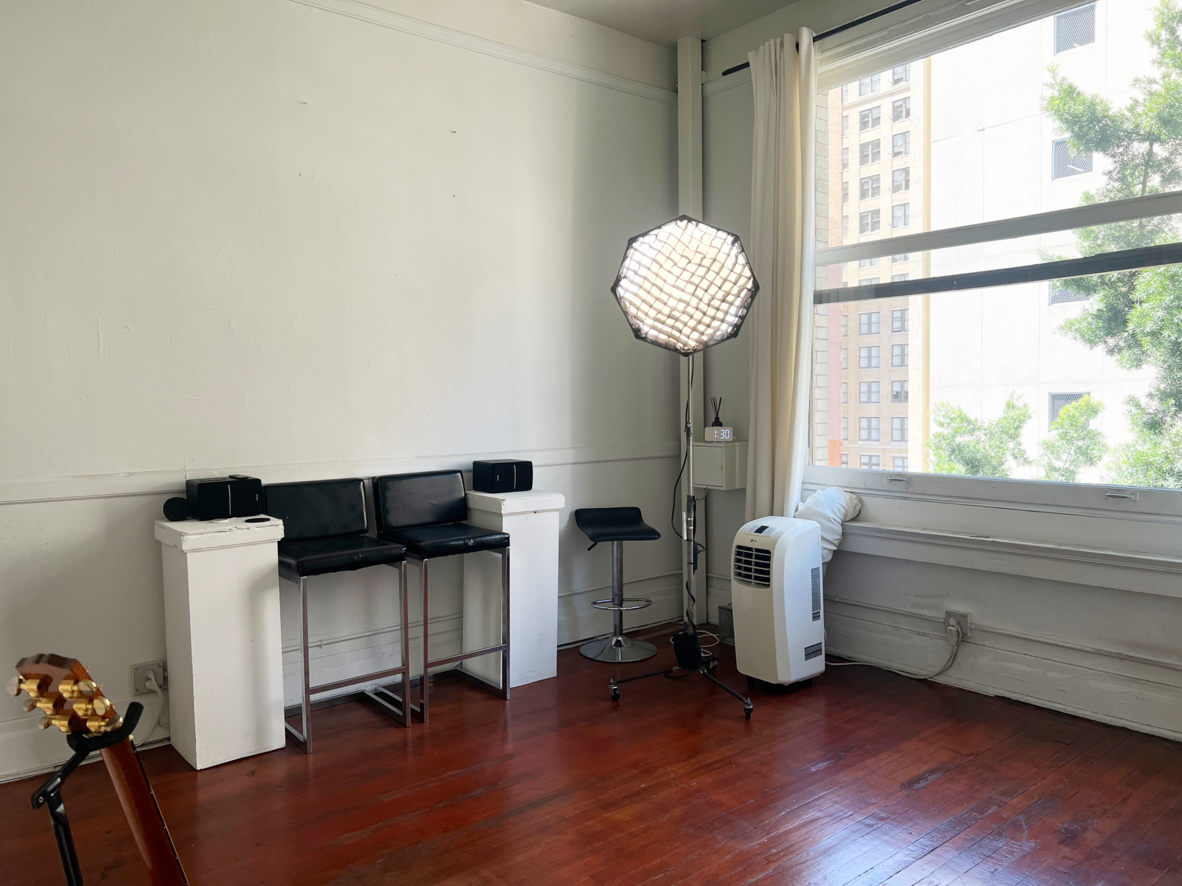 The image shows a minimalistic room with wooden flooring, featuring a stool, a couple of black chairs, a softbox light, an air conditioning unit, and large windows letting in natural light.