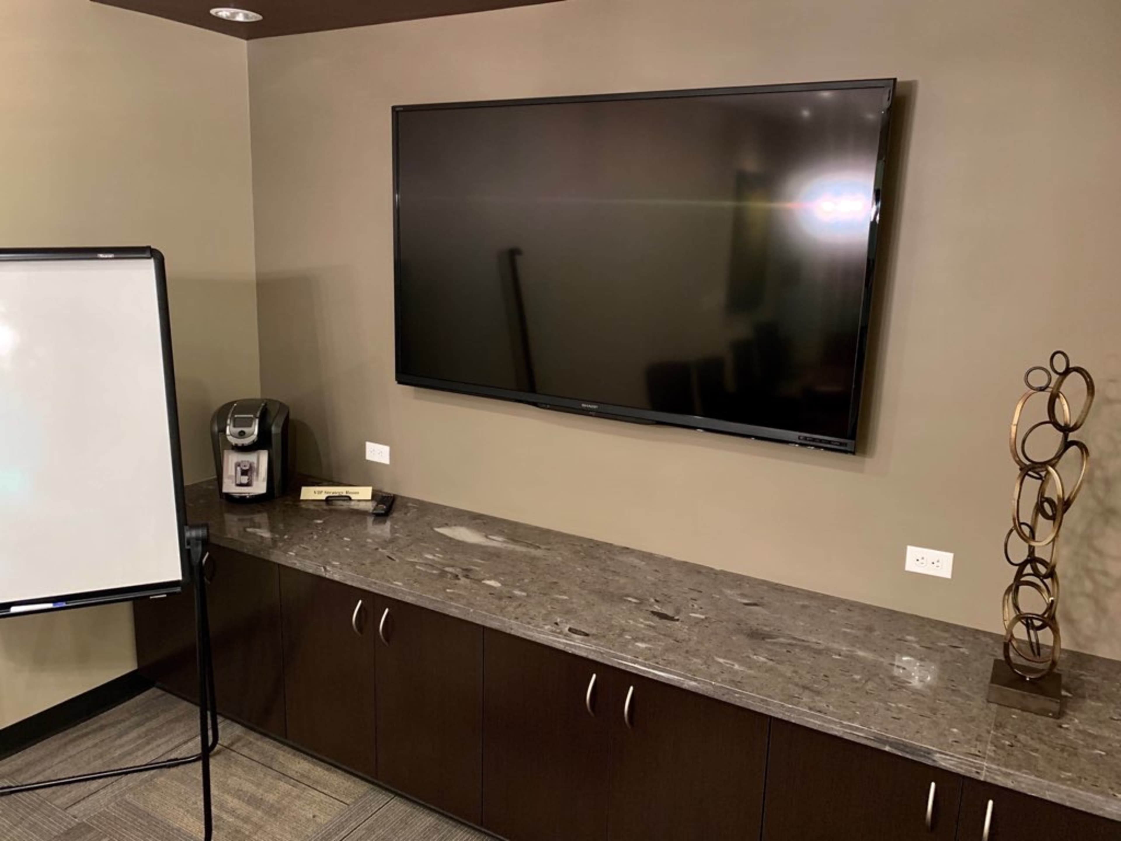 The image shows a conference room with a large flat-screen TV mounted on the wall above a dark cabinet, alongside a whiteboard and a coffee maker.
