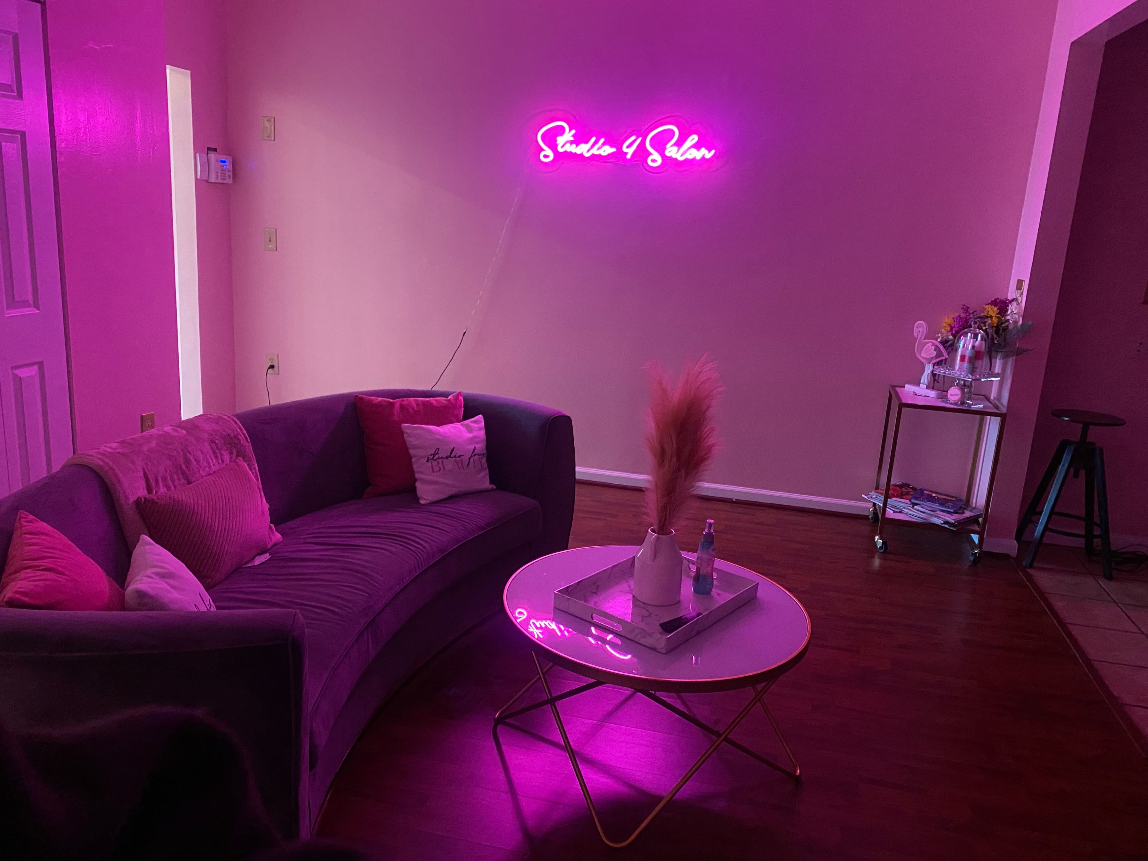 A stylish lounge area with a curved gray couch, a round coffee table, and a neon sign reading "Studio 4 Salon," all in a pink-hued room.