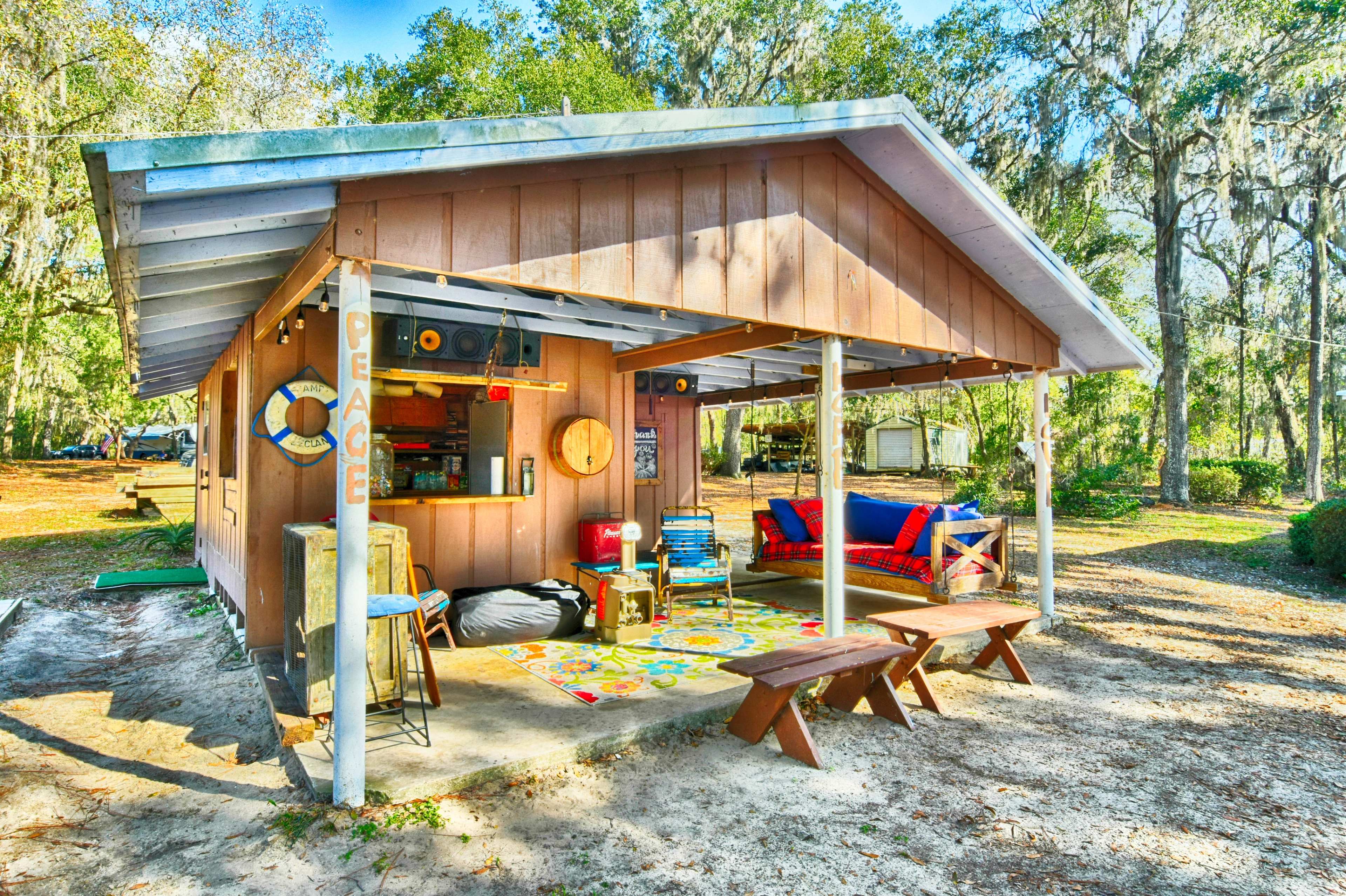 A small wooden cabin with a front porch is set in a wooded area, featuring colorful furnishings and a picnic table outside.