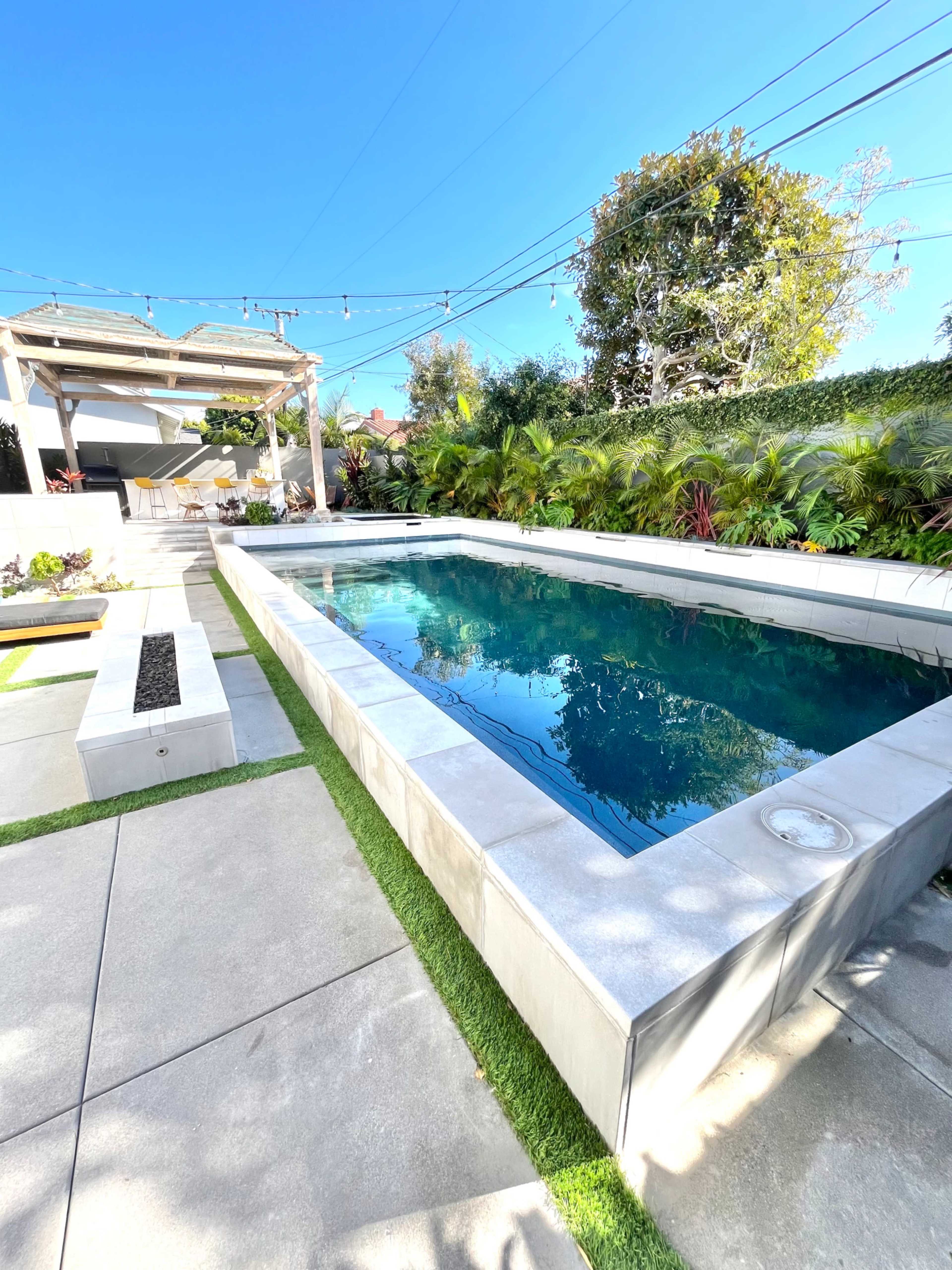 The image shows a rectangular swimming pool surrounded by concrete paving and greenery, with a pergola and outdoor seating area in the background.