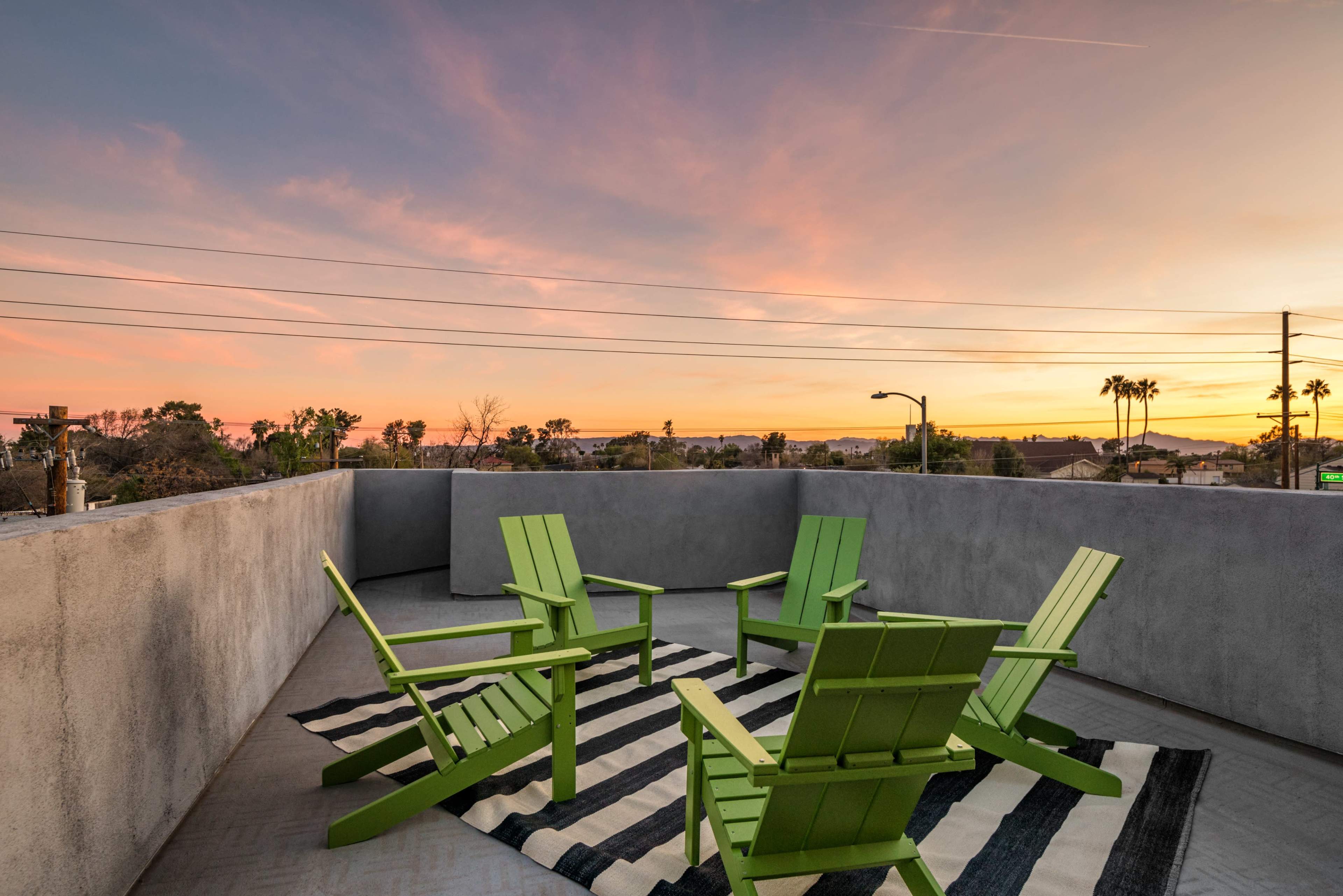 A rooftop terrace features four green chairs arranged around a striped rug, with a colorful sunset in the background.